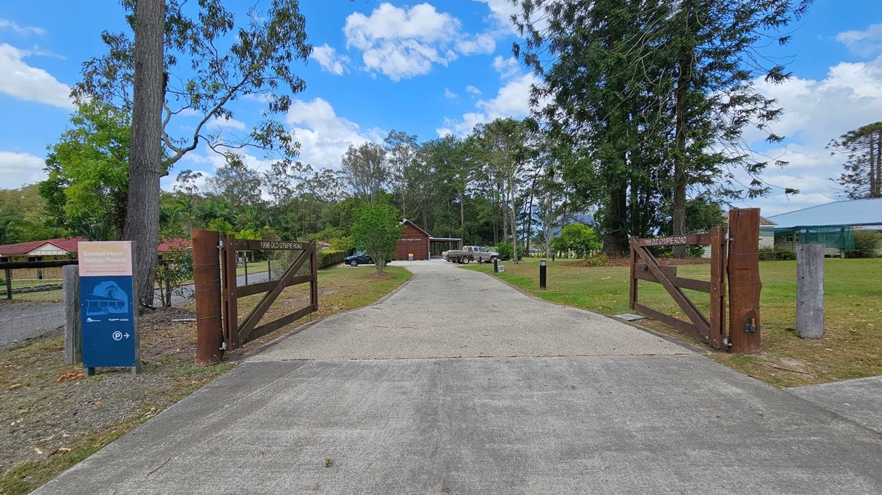 A paved path leads to a wooden gate with a sign reading "Bankfoot House Heritage Precinct" on the left. Tall trees and a small building are visible in the background. A parking sign with an arrow is on the right, next to a road. A grassy area with scattered leaves surrounds the path, and a utility pole stands nearby.