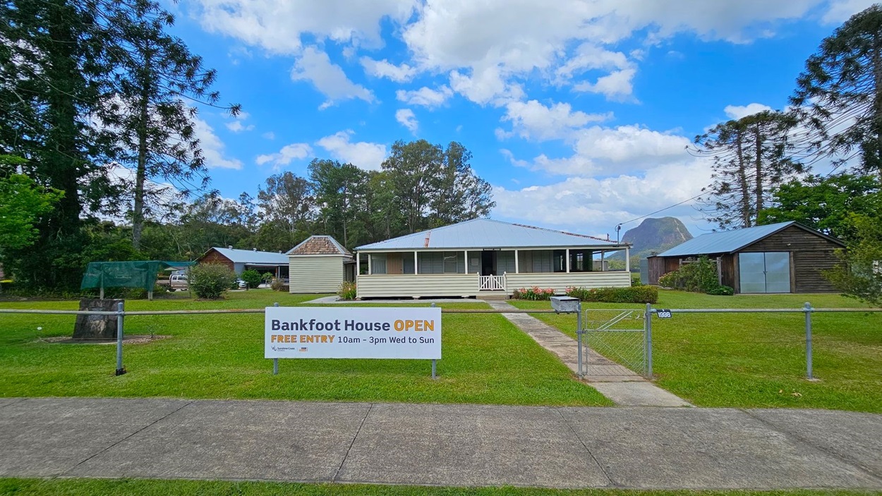 A single-storey house with a corrugated metal roof is in the centre, surrounded by a well-maintained lawn. To the left, there is a small wooden structure and a large tree. On the right, another wooden building with a metal roof is visible. A sign in the foreground reads "Bankfoot House OPEN" with visiting hours. A concrete path leads to the house, and a metal gate is part of the fence enclosing the property. Trees and a mountain are in the background.
