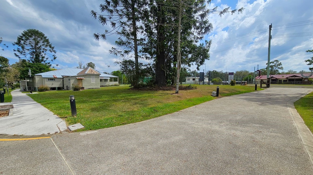A grassy area with a large tree in the centre. On the left, there's a light-coloured house with a metal roof and a concrete path leading towards it. On the right, a paved path curves through the grass, bordered by small black bollards. In the background, there are more houses and utility poles with wires. The sky is partly cloudy.