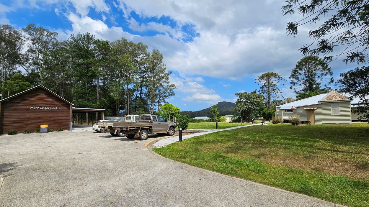 A rural scene with a wooden building on the left labelled "Mary Grigor Centre" surrounded by tall trees. Two utility vehicles are parked nearby. On the right, a small, light-coloured house with a metal roof sits on a grassy area. A path runs between the buildings, and there are trees and a mountain in the background. The ground is a mix of grass and paved surfaces.