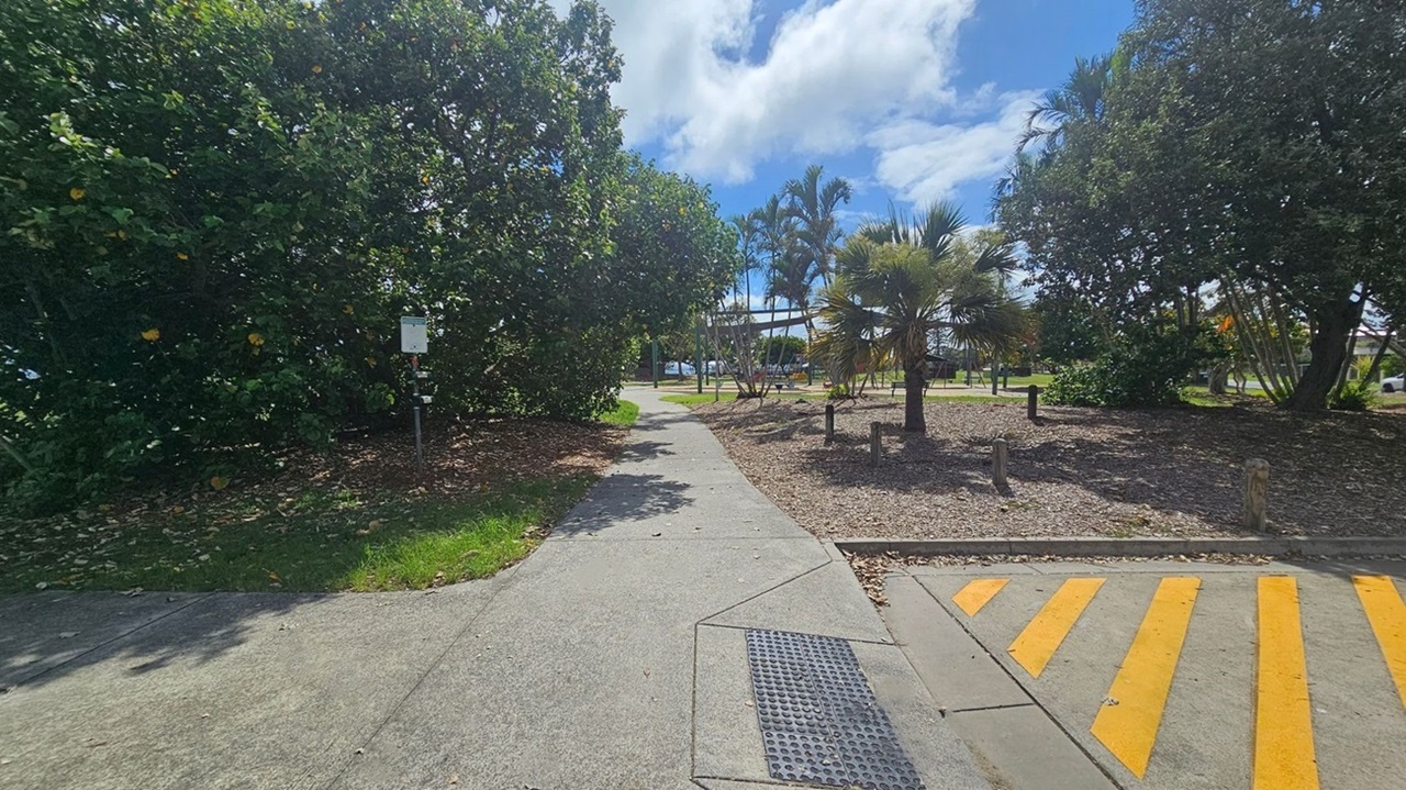 A car park with designated accessible car parking spaces marked by blue symbols on the ground. Yellow lines indicate no parking zones. On the left, a signpost for disabled parking is visible. In the background, there are green bushes and trees, with a black vehicle parked nearby. A white car is parked on the right. The ground is paved with concrete, and there are stone barriers along the grassy area.