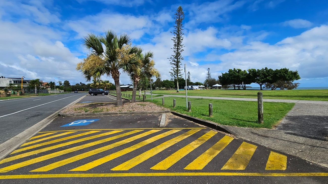 A road with a parking area on the left, featuring a yellow-striped disabled parking space. Several palm trees are planted along the edge of the parking area. In the background, there is a grassy field with a few tall trees and a path running through it. A few cars are parked, and there are some buildings visible in the distance on the left. The sky is partly cloudy.