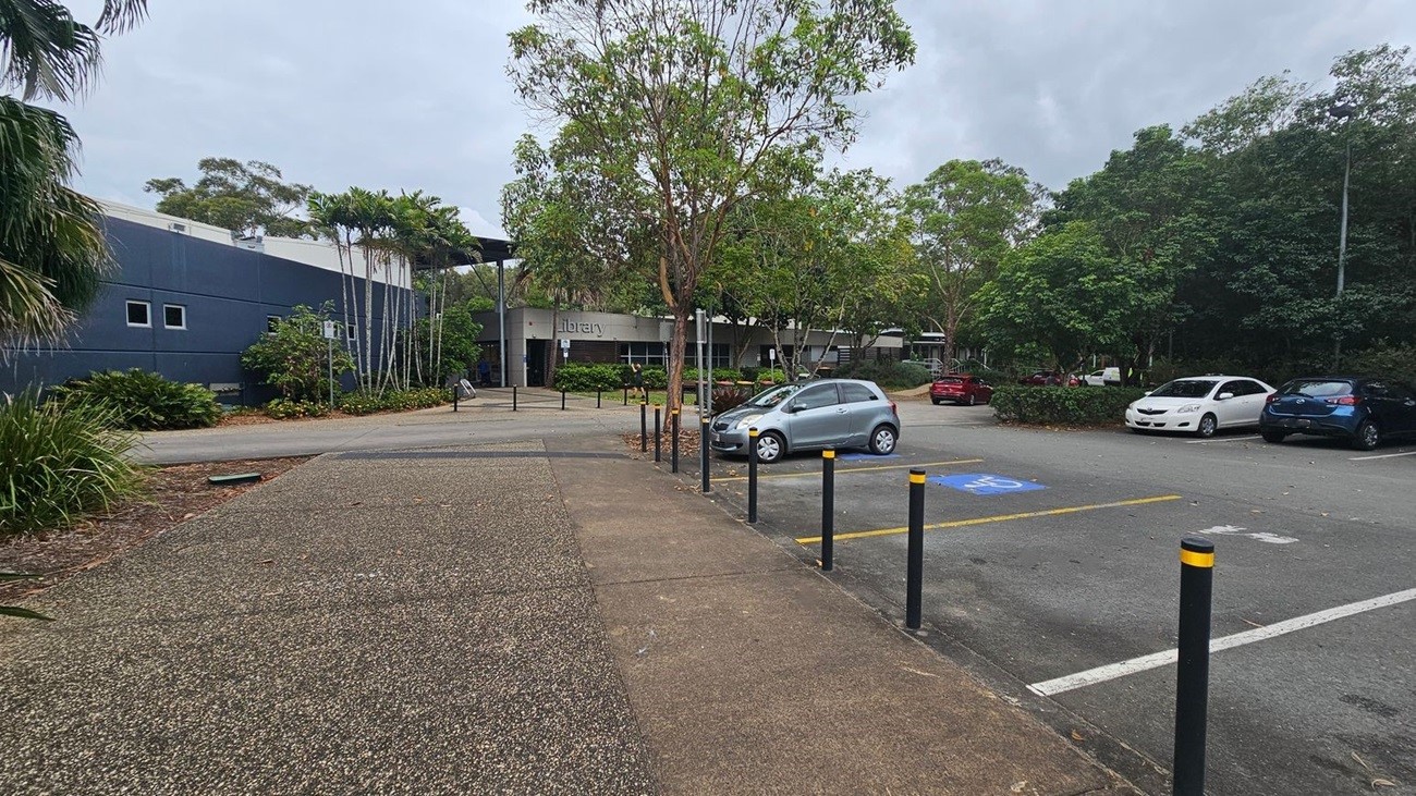 A paved walkway leads to a building labelled "library" in the centre. On the left, there are tall palm trees and a dark grey building with small windows. On the right, a car park with several vehicles, including a silver car in a disabled parking space. The area is surrounded by lush green trees and foliage. Black bollards with yellow tops line the walkway and car park.