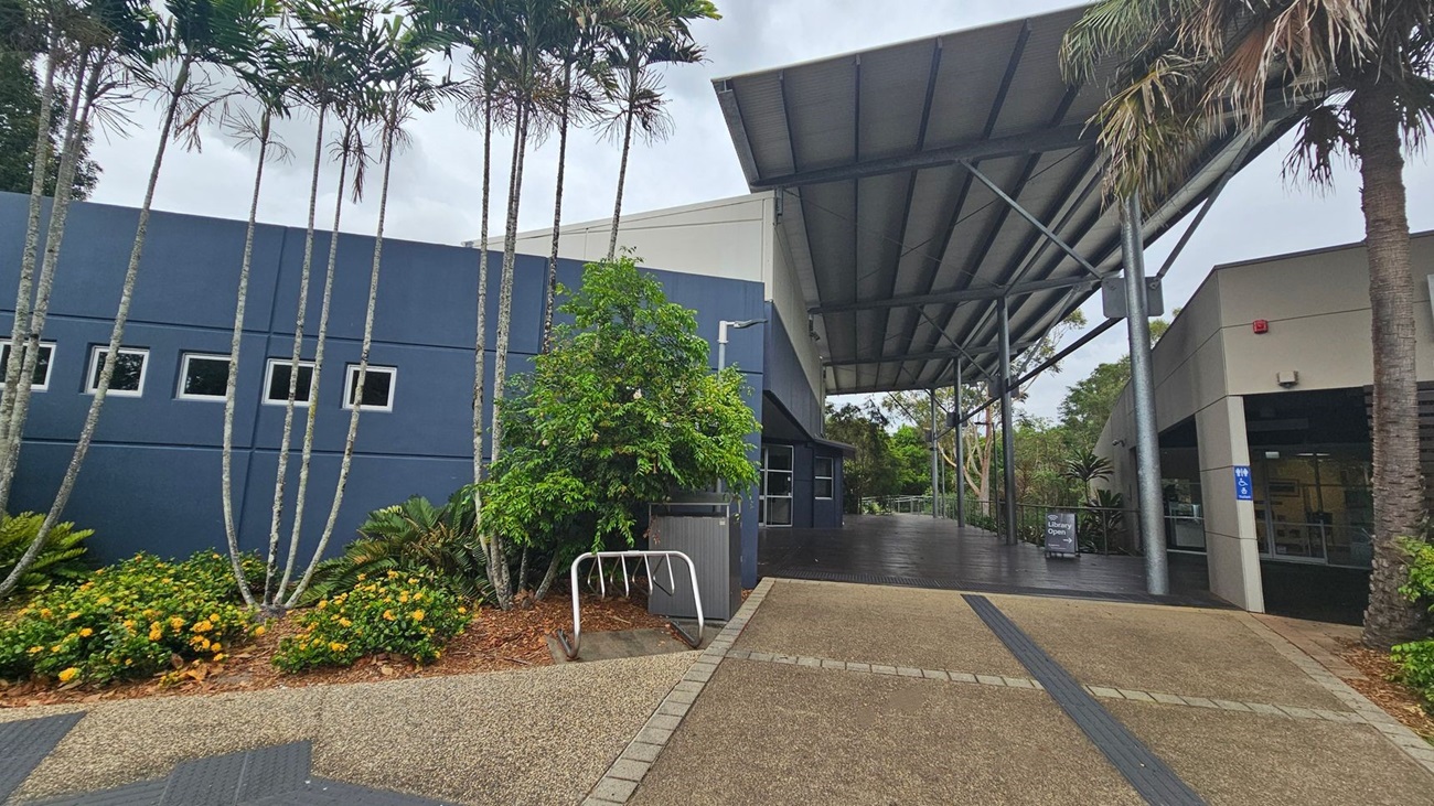 A modern building with a large, overhanging metal roof supported by steel beams. On the left, there is a blue wall with small square windows and tall, slender palm trees in front. Yellow flowers and green shrubs are planted along the base. In the centre, a bike rack is visible. On the right, an entrance with a sign indicating the library is open. The ground is paved with textured tiles, and there are accessible signs near the entrance.