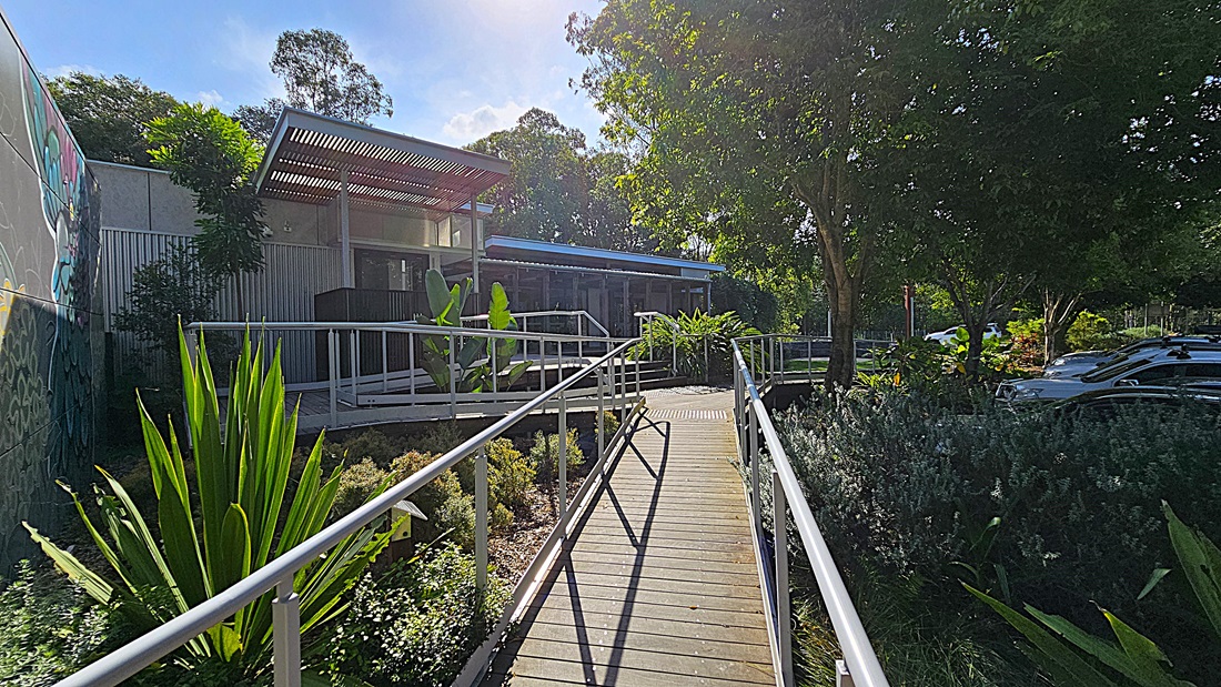 A narrow concrete path leads through a garden towards a modern building on the left. The building has slatted wooden panels and large windows. Dense shrubs and plants line the left side of the pathway, while a red-lidded wheelie bin stands on the right. In the background, trees provide shade, and parked cars are partially visible on a gravel surface. The area is surrounded by lush greenery, contributing to a natural setting.
