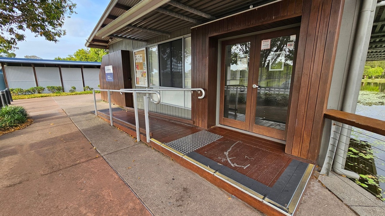 A building with a wooden exterior and glass double doors on the right. A metal handrail runs along a ramp leading to the entrance. The ground is concrete with scattered leaves. On the left, there's a path lined with greenery and a corrugated metal wall in the background. The roof is metal, and a body of water with lily pads is visible on the far right.