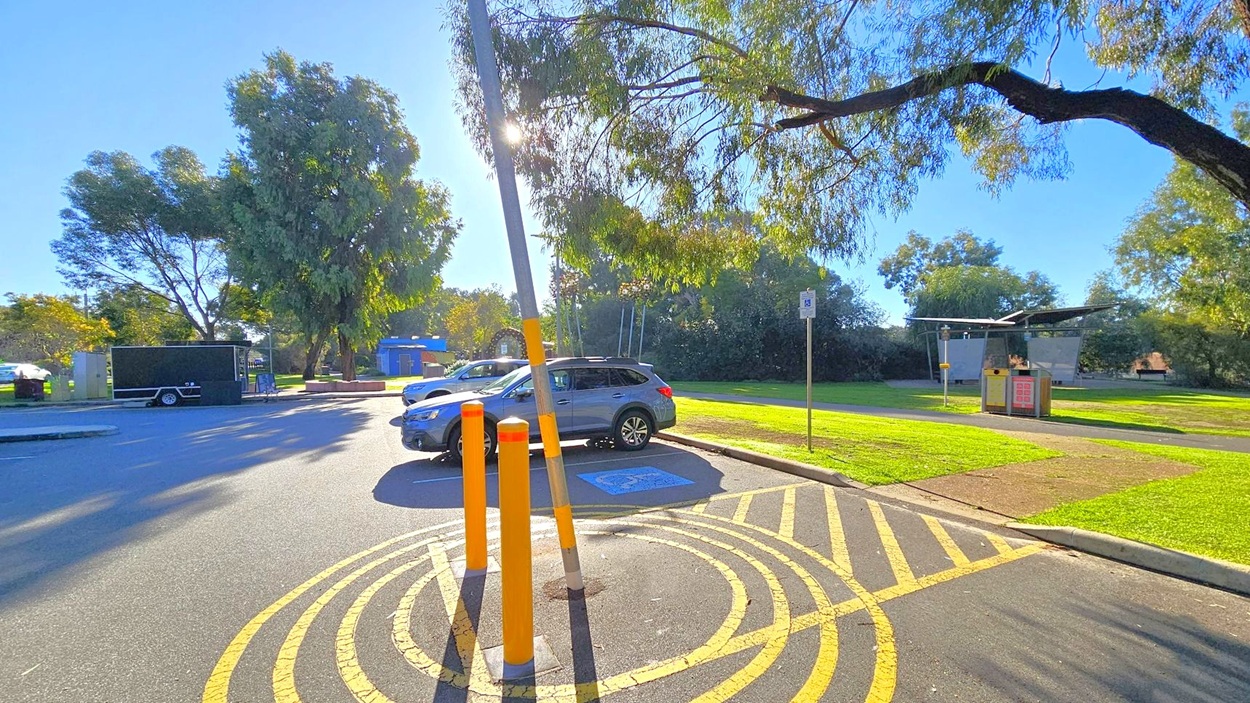 A parking area with a blue car parked in a designated disabled spot, marked by a sign and ground symbol. Three orange bollards and a leaning pole are in the foreground. On the left, a black trailer is parked near large trees. The right side features a grassy area with a small structure, possibly a kiosk or shelter, and more trees in the background. The ground is marked with yellow lines, and sunlight filters through the tree branches.