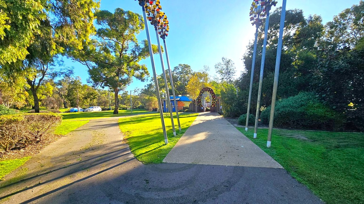 A park scene with a paved path in the centre leading to an archway adorned with colourful decorations. On the left, there are tall trees and a few parked cars visible in the distance. On the right, more trees and bushes create a dense green area. Several tall poles with decorative elements are positioned near the path. The grass is neatly trimmed, and the area is well-lit by sunlight.