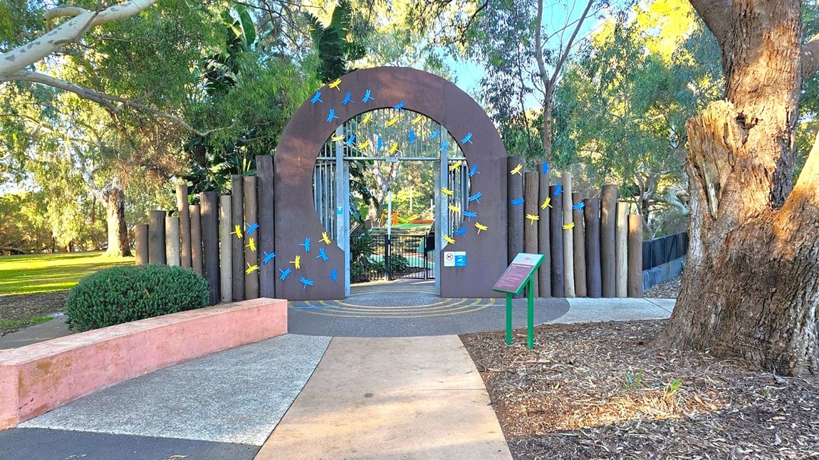 A large, circular archway decorated with blue and yellow dragonfly motifs stands in the centre, leading to a gated entrance. The arch is flanked by vertical wooden posts. On the left, there's a curved pink concrete bench and a bush. On the right, a large tree trunk and a green signpost with a slanted information board. The ground is a mix of concrete and mulch, with trees and greenery in the background.