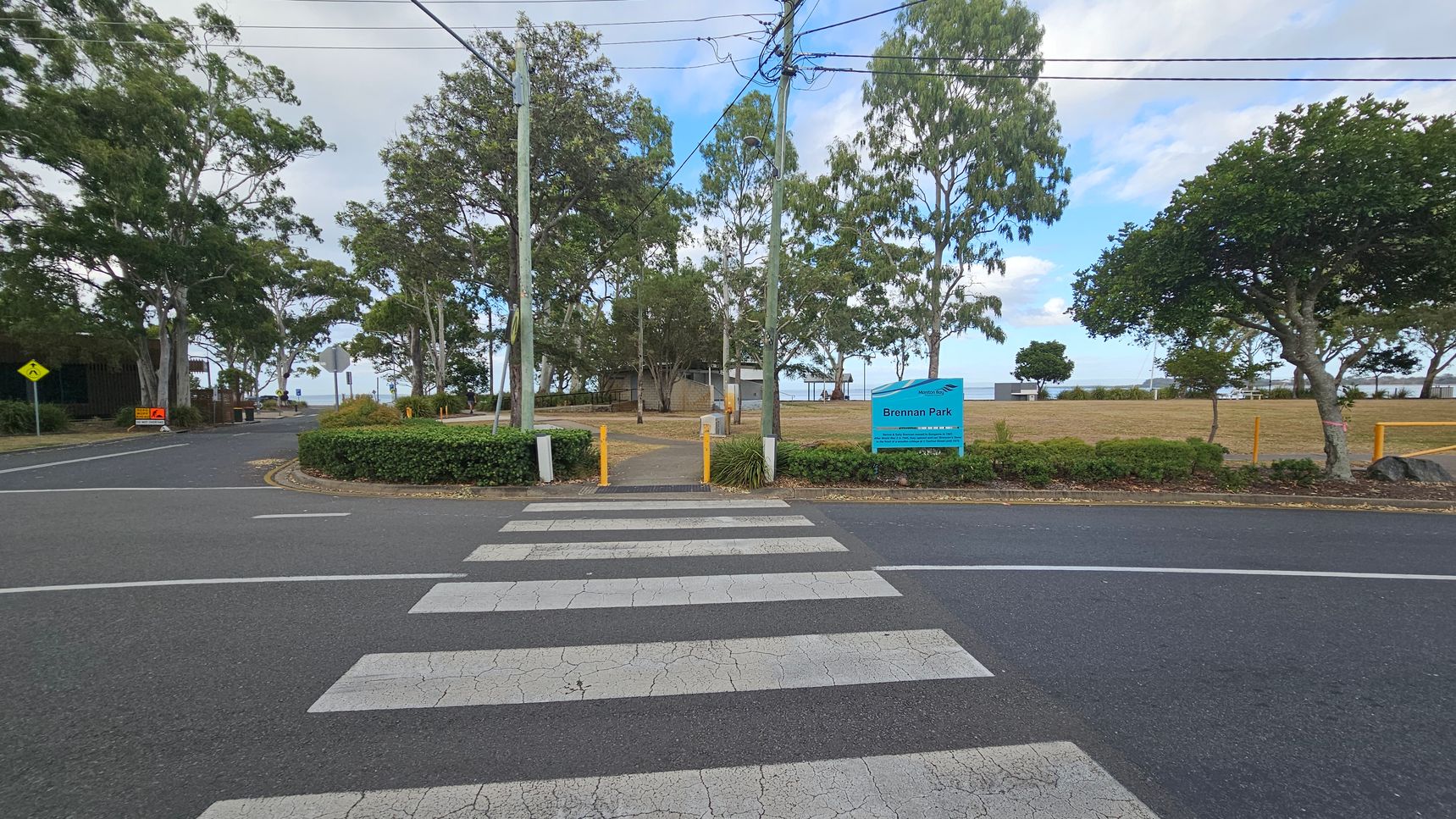 A zebra crossing leads to a park entrance with a blue sign reading "Brennan Park" on the right. The park is bordered by trees and has a grassy area. Yellow bollards and a path are visible beyond the crossing. Power lines run overhead, and there are more trees and a road to the left. The background shows a glimpse of water beyond the park.
