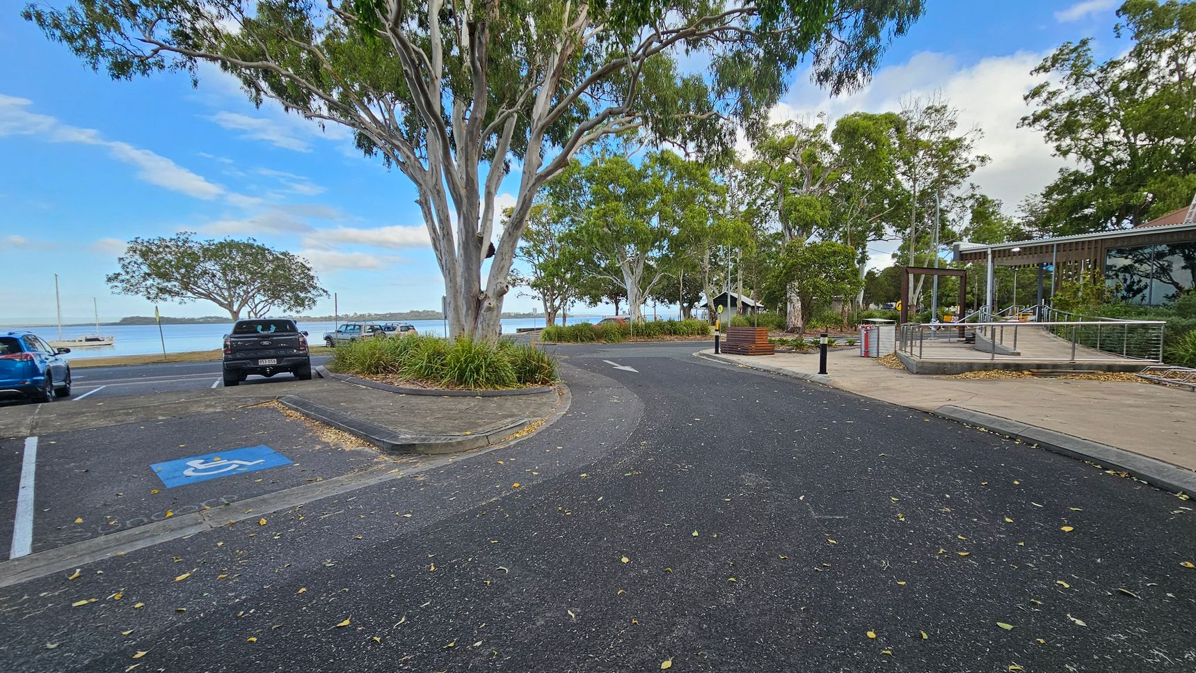 A paved road curves through a park-like area with large trees on both sides. On the left, there are parked cars, including a blue vehicle and a black one in a disabled parking space. In the background, a body of water with a boat is visible. On the right, a modern building with glass windows and a ramp is surrounded by landscaped greenery. The ground is covered with scattered leaves.