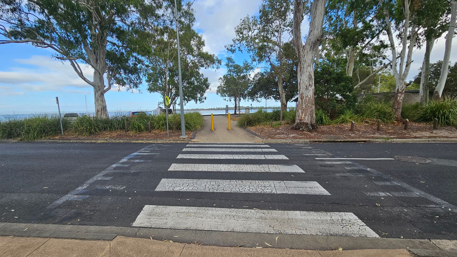 A zebra crossing leads to a path flanked by two yellow bollards. Large trees with thick trunks and green foliage are on both sides, with grass and mulch at their bases. Beyond the path, a body of water is visible in the distance. The road is dark with visible wear, and the pavement is light brown. A metal pole with a sign is on the left side of the path.