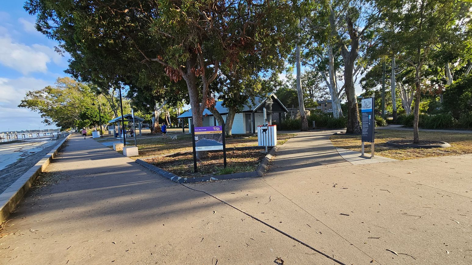 A paved walkway runs along the left side, bordered by a low stone wall and a body of water. Trees line the path, providing shade. In the centre, a signpost and a small building with a sloped roof are visible. On the right, another signpost stands near a grassy area with more trees. A person walks a dog in the distance. The ground is covered with scattered leaves and the area is well-lit by natural light.