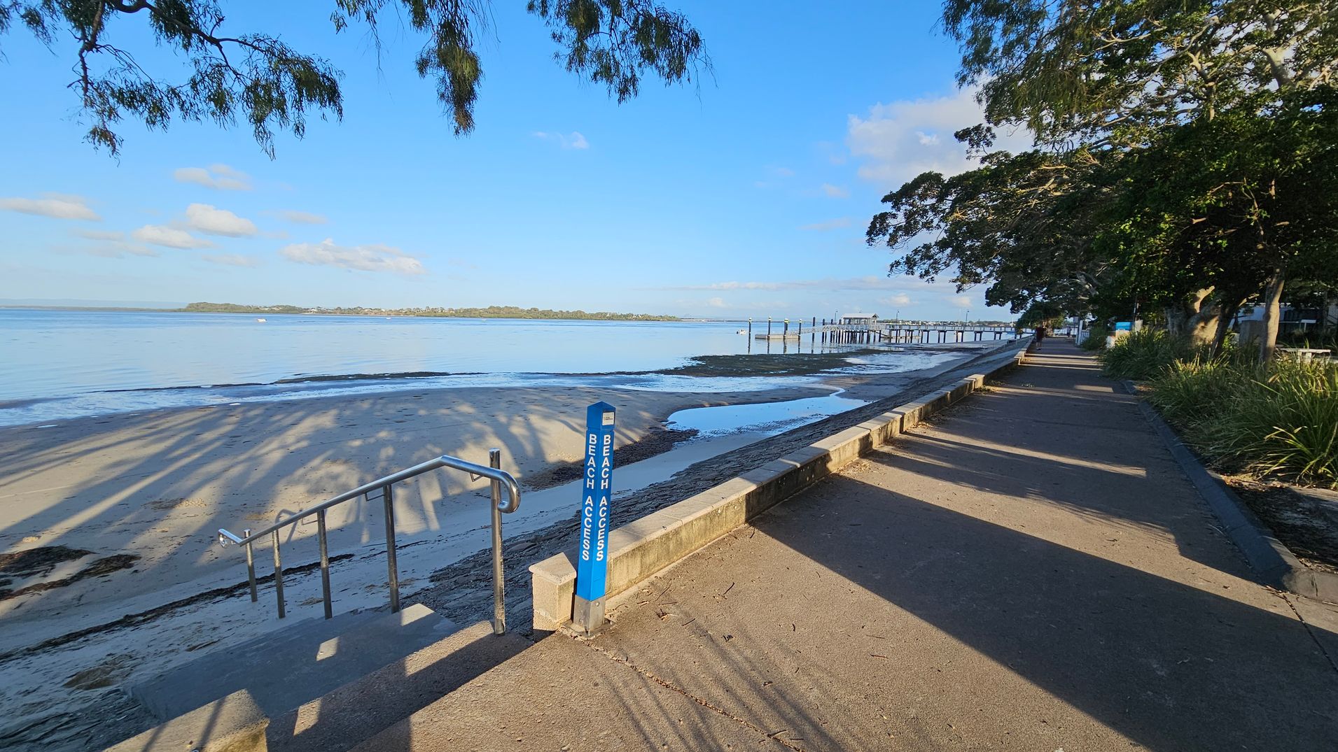 A coastal scene with a sandy beach on the left, leading to calm blue water. A metal railing provides access to the beach, marked by a blue "Beach Access" sign. On the right, a paved walkway is lined with trees casting long shadows. In the distance, a pier extends into the water. The background features a clear sky with a few clouds and a distant shoreline.