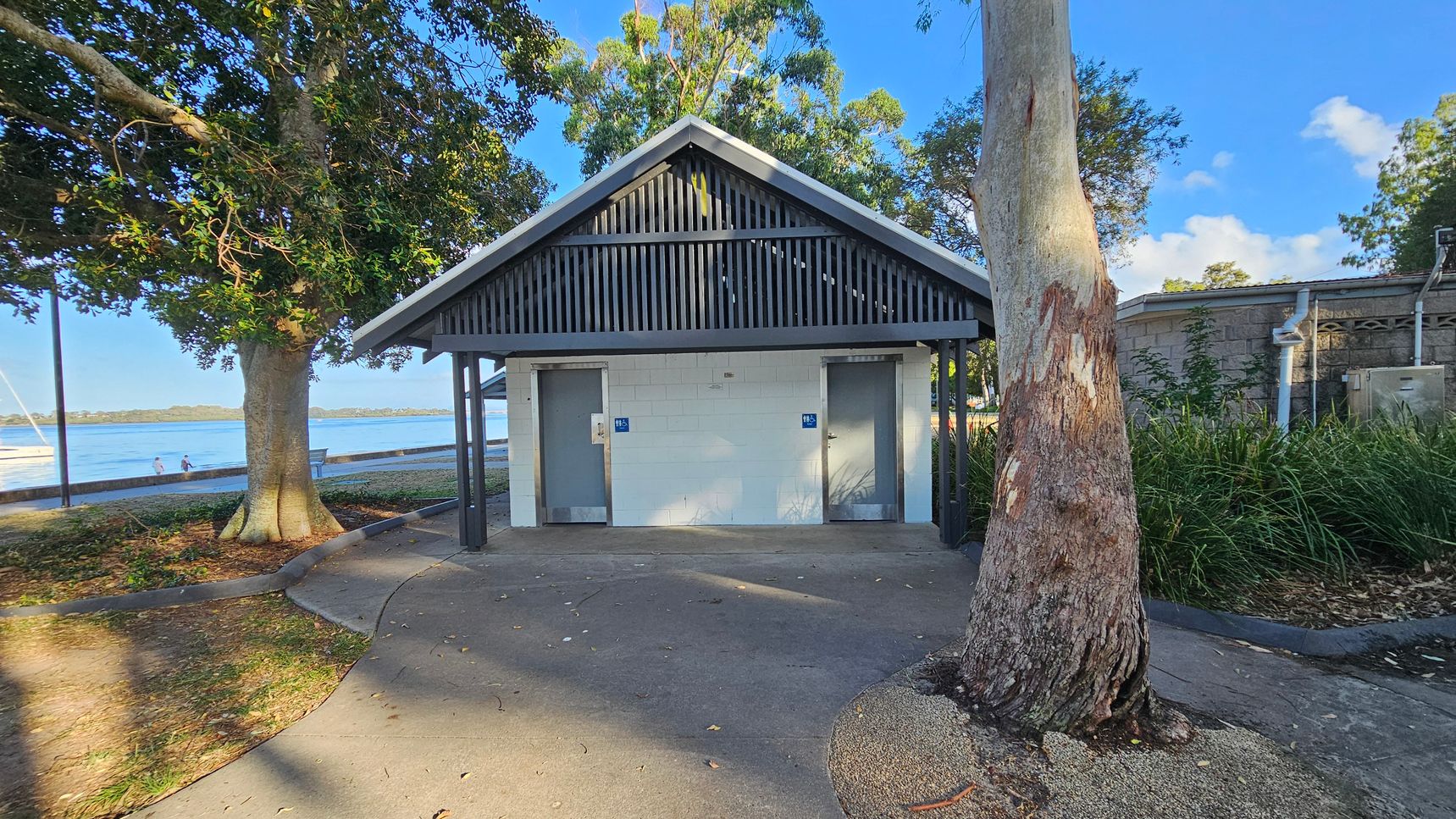 A small public restroom building with a gabled roof and two doors, each marked with a blue sign. The building is surrounded by trees, with a large tree trunk in the foreground on the right. To the left, a path leads towards a waterfront area with a couple of people sitting on a bench near the water. The ground is a mix of concrete and natural earth, and there is a grassy area with plants on the right side.