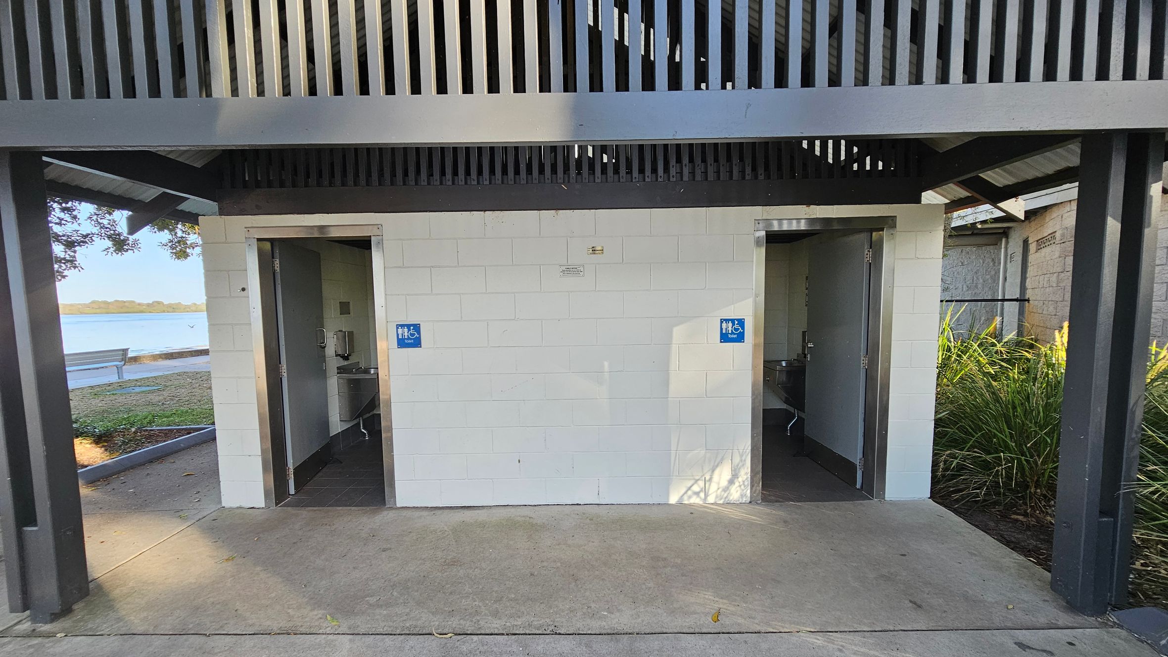 Two public toilets with open doors are visible, each marked with a blue sign indicating accessibility. The structure is made of white bricks with a grey metal roof featuring vertical slats. On the left, a bench and a body of water are visible in the background. On the right, there are green plants. The floor is concrete, and the ceiling is not visible.