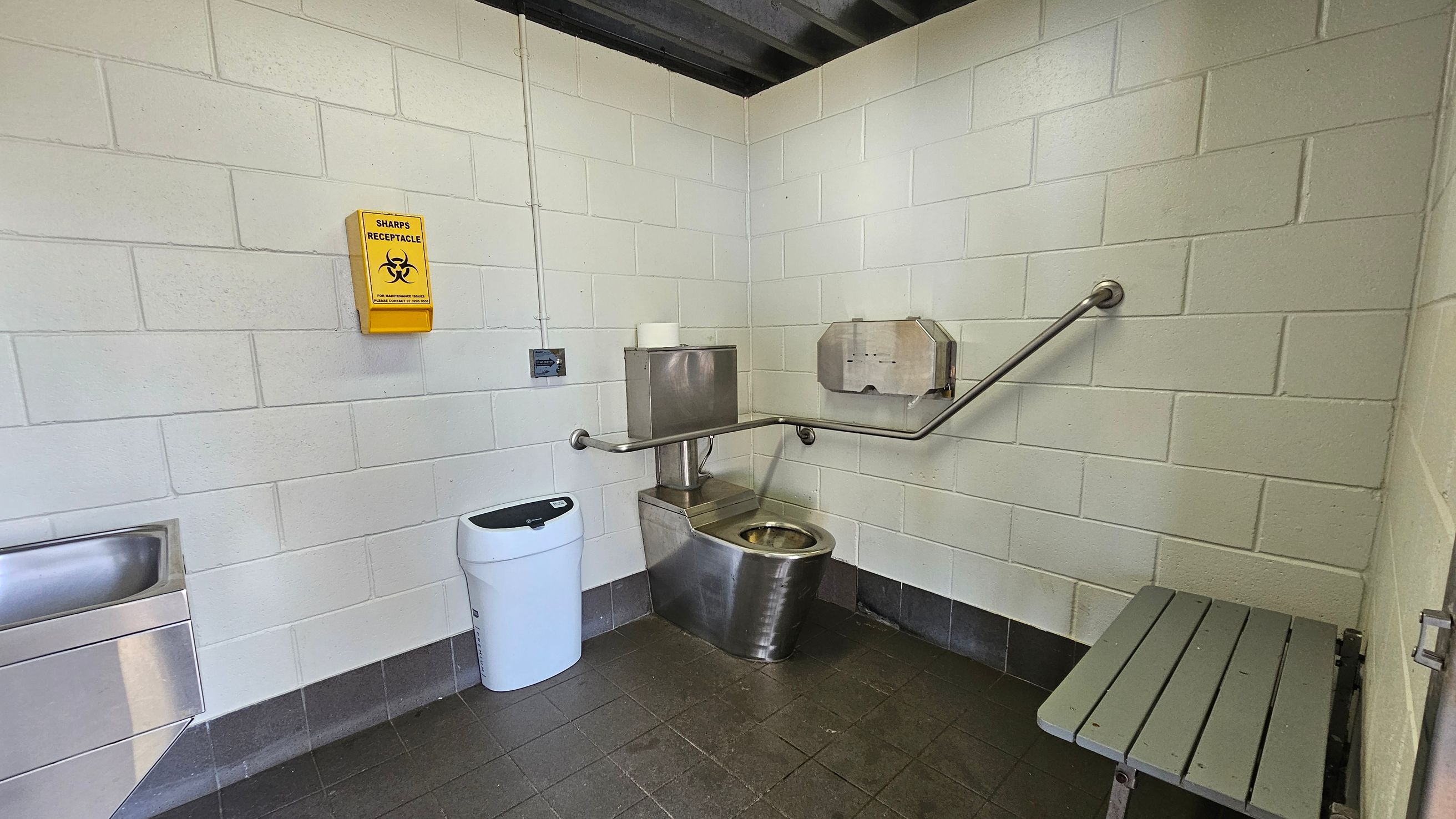 A public restroom with white tiled walls and a dark tiled floor. On the left, a stainless steel sink is mounted on the wall. In the centre, a stainless steel toilet with a roll of toilet paper on top is accompanied by a wall-mounted paper dispenser and a grab bar. A yellow sharps receptacle is fixed to the wall above. A white bin is positioned near the toilet. On the right, a grey bench is attached to the wall. The ceiling is dark with visible beams.