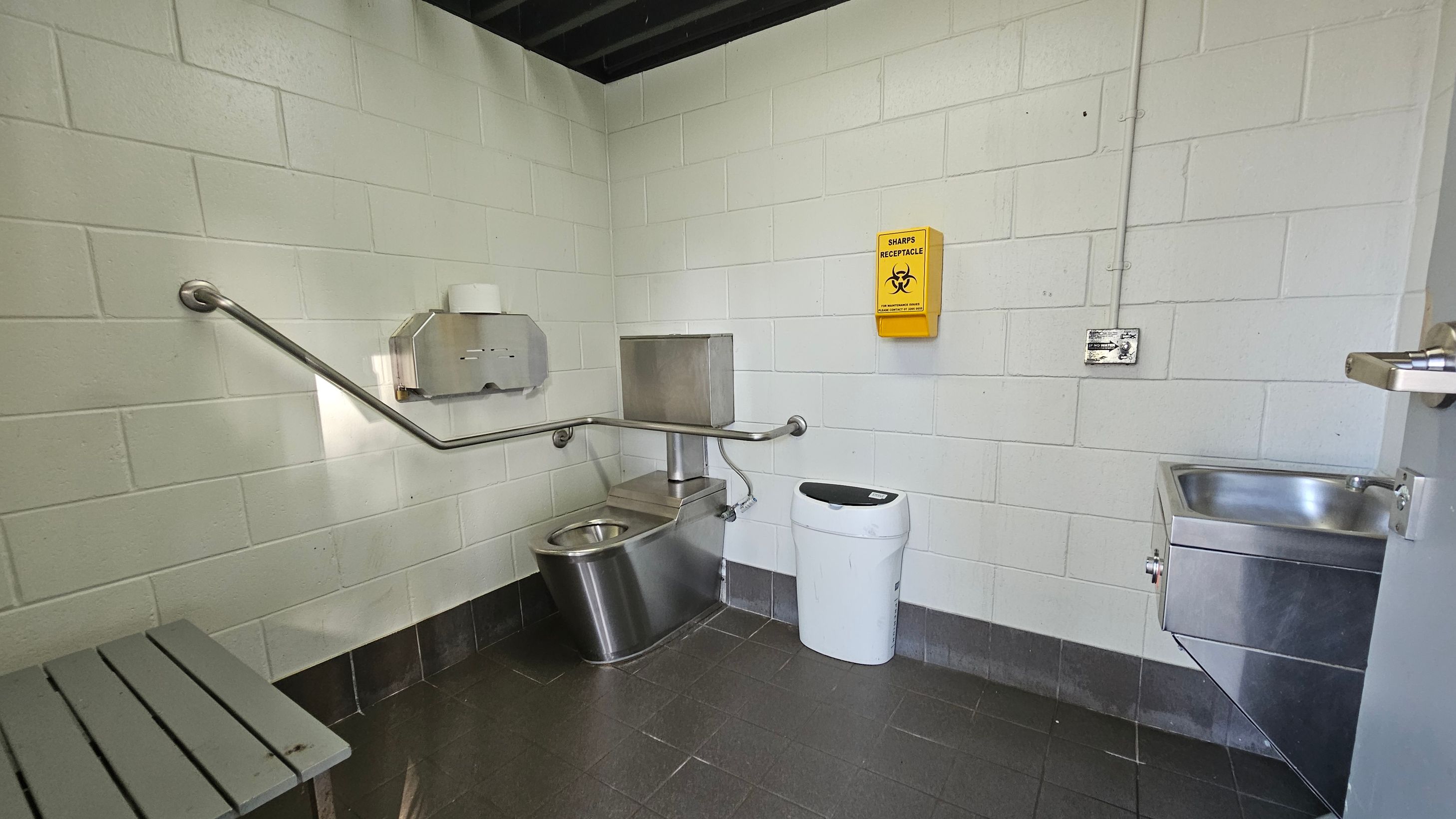 A small, utilitarian bathroom with grey tiled flooring and white brick walls. On the left, a metal bench is visible. In the centre, a stainless steel toilet with a wall-mounted toilet paper dispenser and a grab bar is present. Above the toilet, a yellow sharps receptacle is mounted on the wall. To the right, a small stainless steel sink is attached to the wall, with a door partially open next to it. The ceiling is dark with exposed beams.