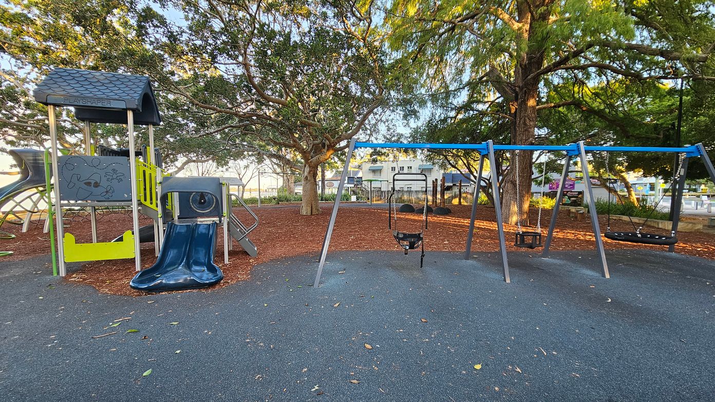 A playground with a play structure on the left featuring a small roof, climbing elements, and a blue slide. To the right, there is a swing set with multiple swings. The ground is covered with a dark rubber surface, and the area is surrounded by trees with dense foliage. In the background, there are buildings and a fence, with some benches and signs visible. The scene is well-lit, suggesting daylight.