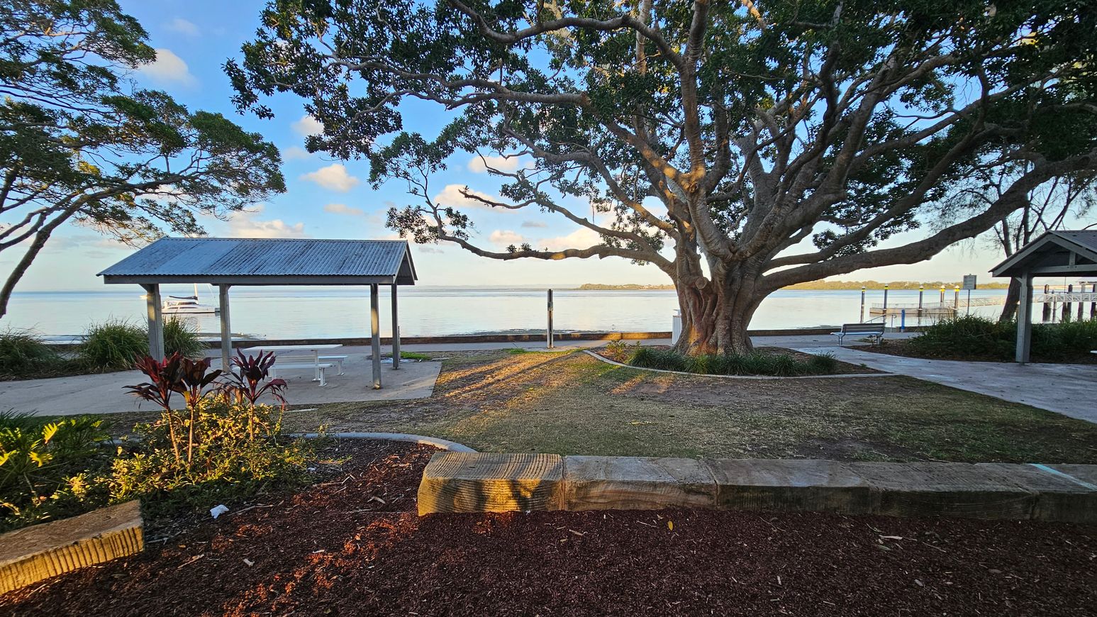 A large tree with sprawling branches dominates the centre, casting shadows on the ground. To the left, a small shelter with a corrugated metal roof stands near the water's edge. On the right, another similar shelter is partially visible. The foreground features a landscaped area with red mulch and plants. The background shows a calm body of water with a boat and a distant shoreline. The ground is a mix of grass and paved paths.