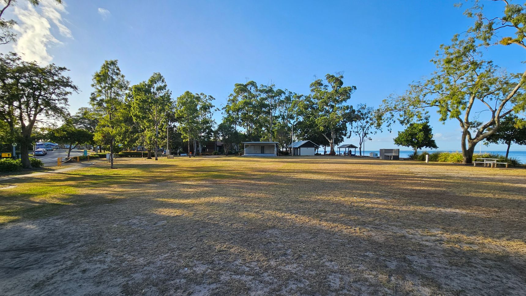 A grassy park area with patches of dry grass in the foreground. In the middle, there are several trees providing shade, and a few small structures, possibly shelters or kiosks. To the left, a road with a car and more trees. On the right, a picnic table and a view of the water in the background. The sky is clear and blue.