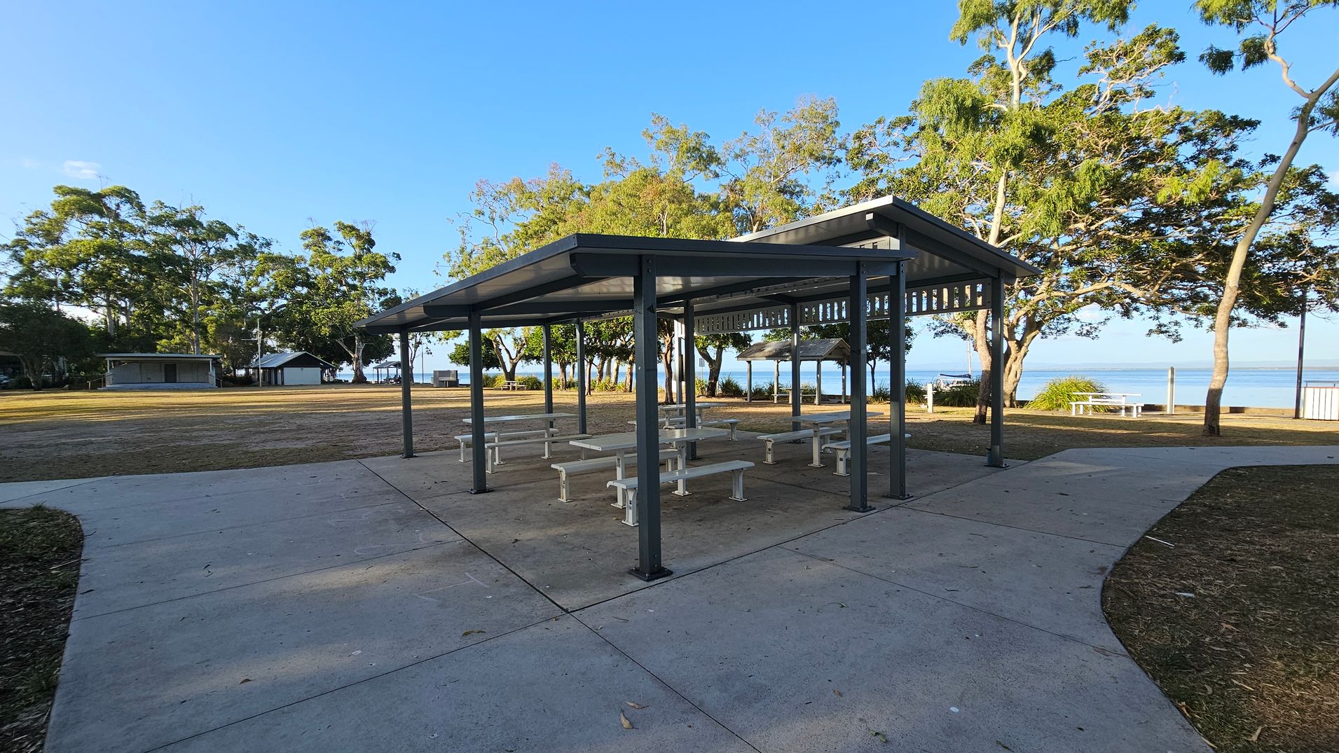 A covered picnic area with several white benches and tables is in the centre. The structure has a metal roof supported by dark-coloured poles. Surrounding the area is a concrete path with patches of grass and mulch. In the background, there are trees and a glimpse of water, suggesting a coastal location. To the left, there are small buildings, possibly restrooms or storage facilities. The area is well-maintained and open.