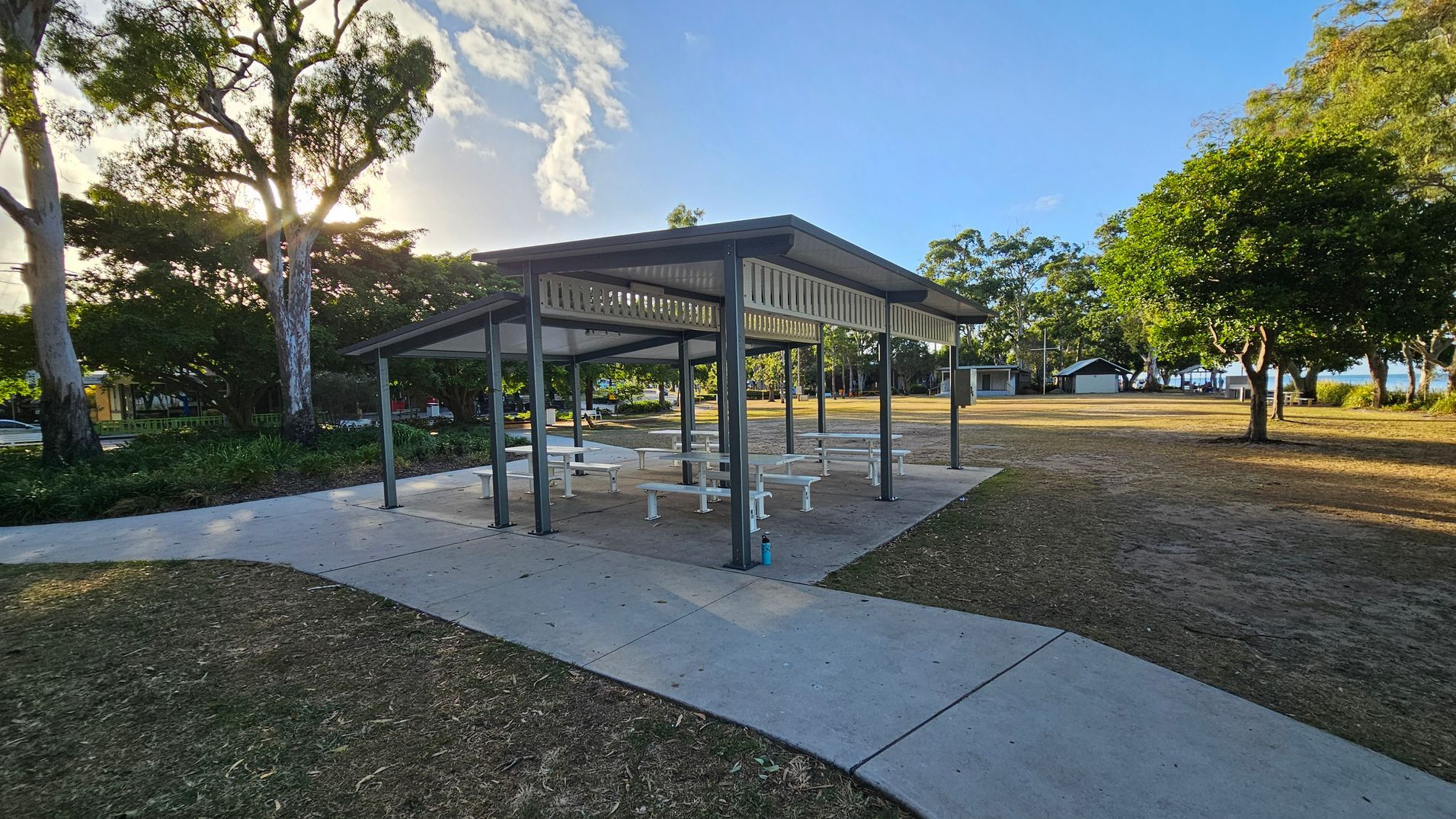 A park setting with a covered picnic area in the centre, featuring several white tables and benches. The structure has a metal roof supported by dark-coloured posts. On the left, tall trees with dense foliage partially block the sun. On the right, more trees and open grassy areas are visible, with a small building in the distance. The ground is a mix of concrete paths and dry grass.