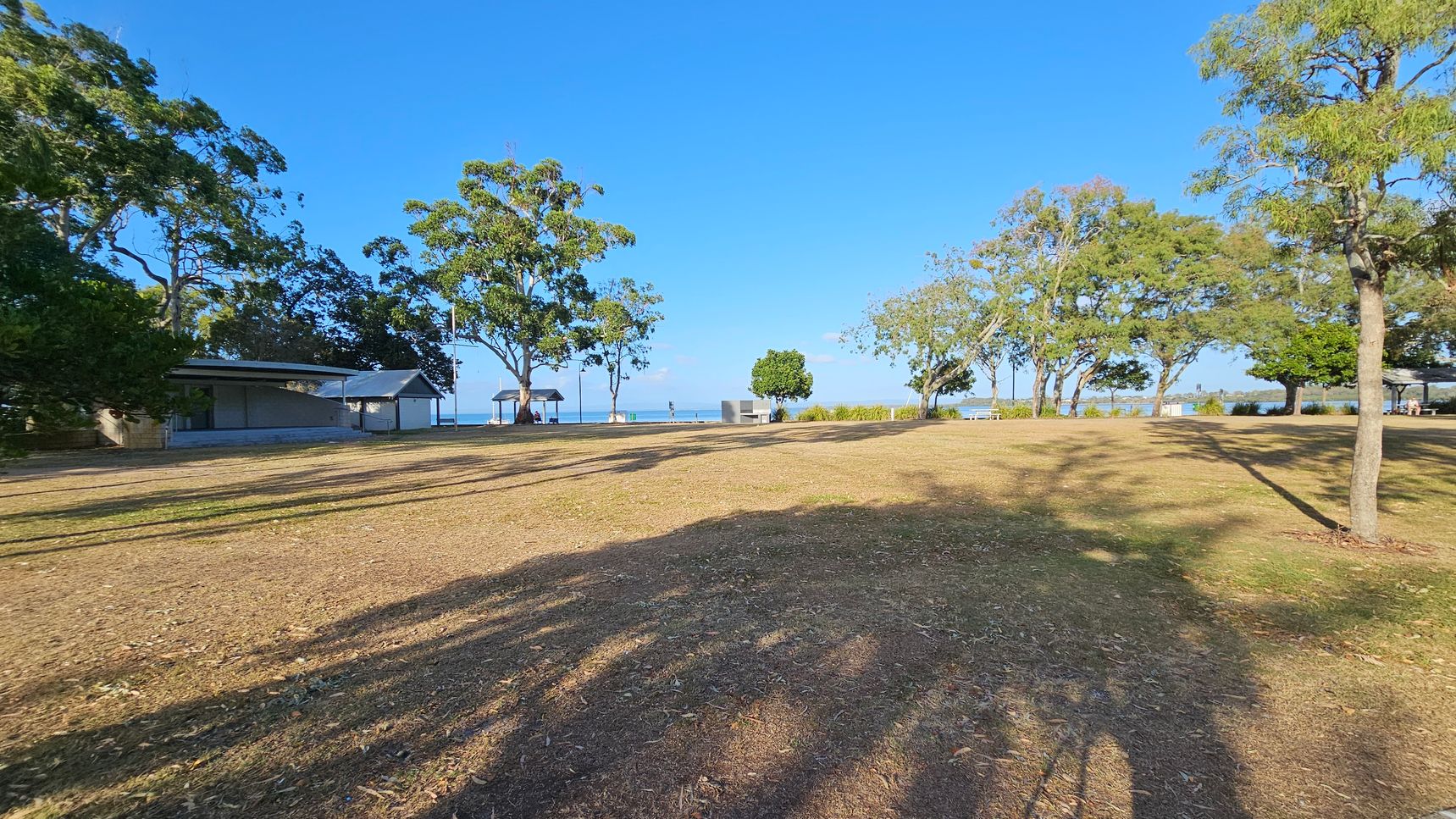 A grassy area with scattered trees casting long shadows. On the left, there is a small building with a sloped roof, partially obscured by trees. In the centre, a path leads towards the water, with a few people visible near a gazebo. On the right, more trees line the edge of the grass, with a glimpse of the sea in the background. The ground is a mix of dry grass and patches of dirt.