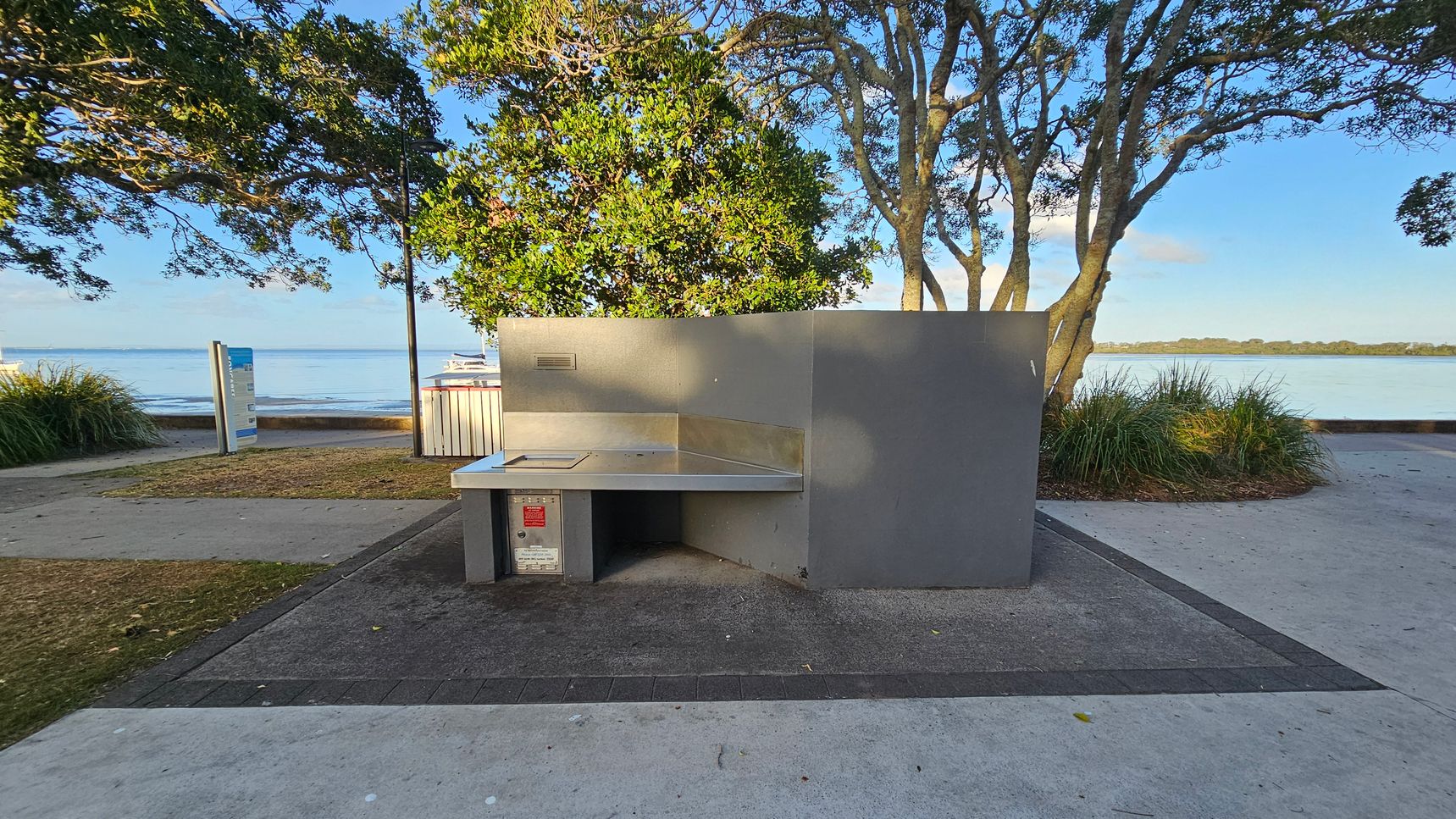 A public barbecue area with a stainless steel cooking surface is in the foreground, set on a paved area. To the left, a path leads towards a sign and a grassy patch. In the background, there are trees with dense foliage and a view of a calm body of water. A few boats are visible on the water, and there is a low wall separating the path from the water's edge. The scene is well-lit, suggesting daylight.