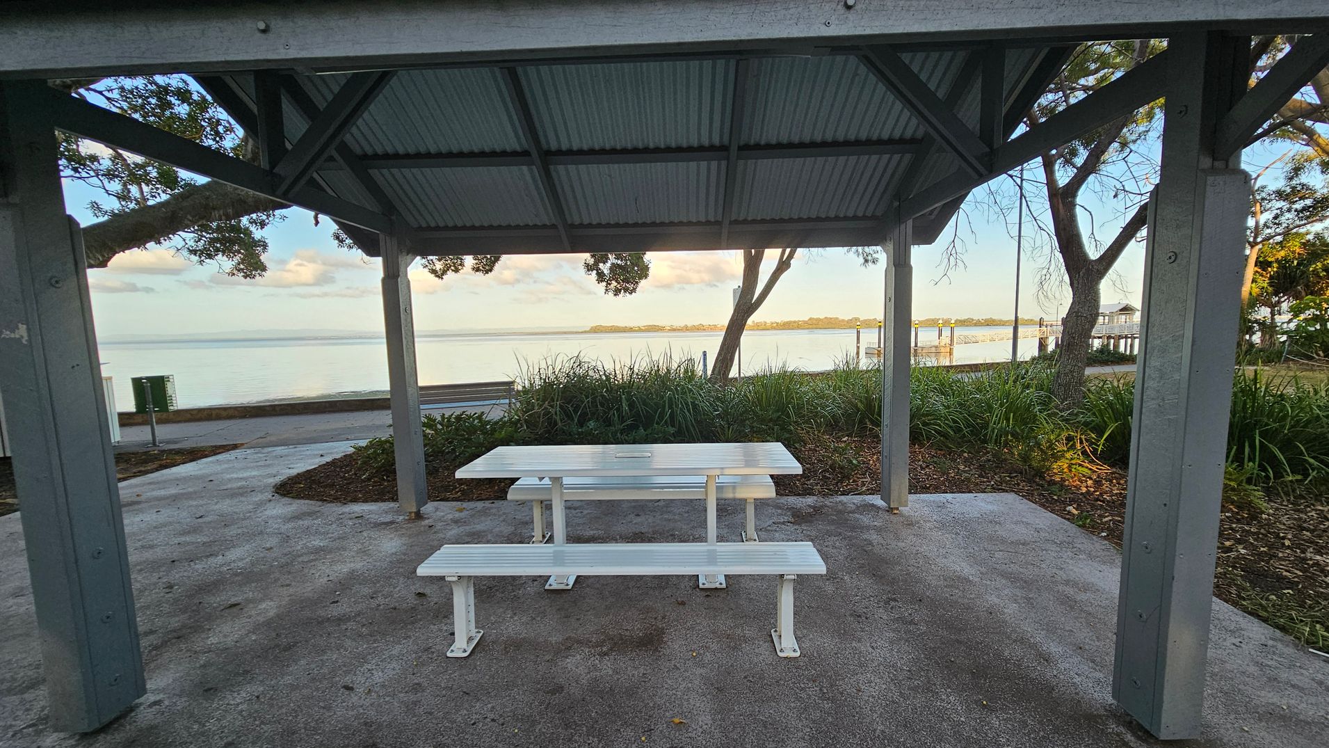 A sheltered picnic area with a white table and benches on a concrete floor. The structure has a corrugated metal roof supported by wooden beams. In the background, there is a view of a calm body of water with a distant shoreline. Trees and greenery surround the area, and a pathway runs parallel to the water. A small pier extends into the water on the right side.