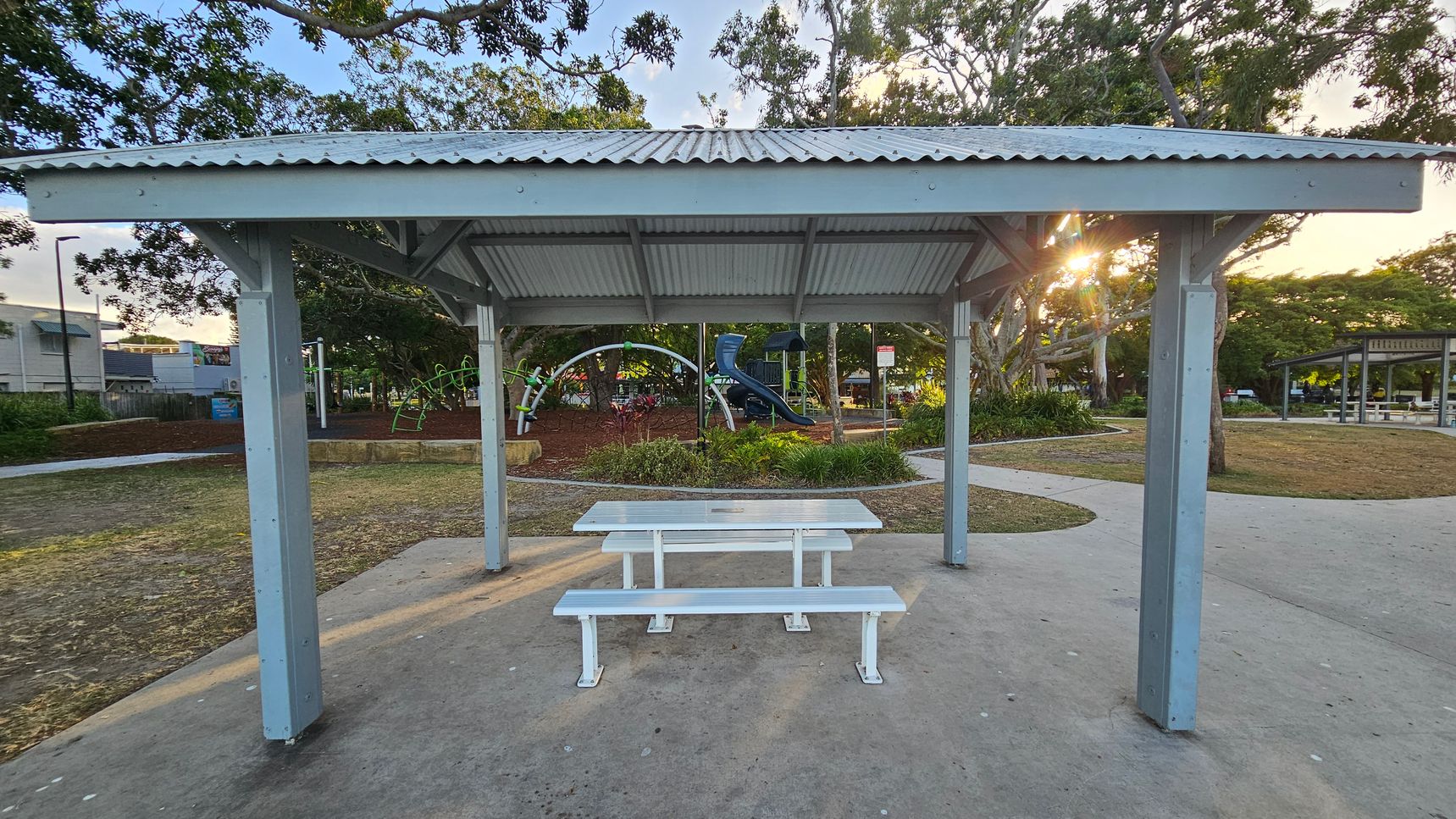 A park setting with a metal shelter in the foreground, featuring a corrugated roof and supported by four posts. Underneath is a white picnic table with benches. In the background, a playground with climbing structures and a slide is visible, surrounded by trees and greenery. A path curves around the area, and another shelter is seen to the right. The ground is a mix of concrete and grass, with some mulch around the playground. Sunlight filters through the trees.