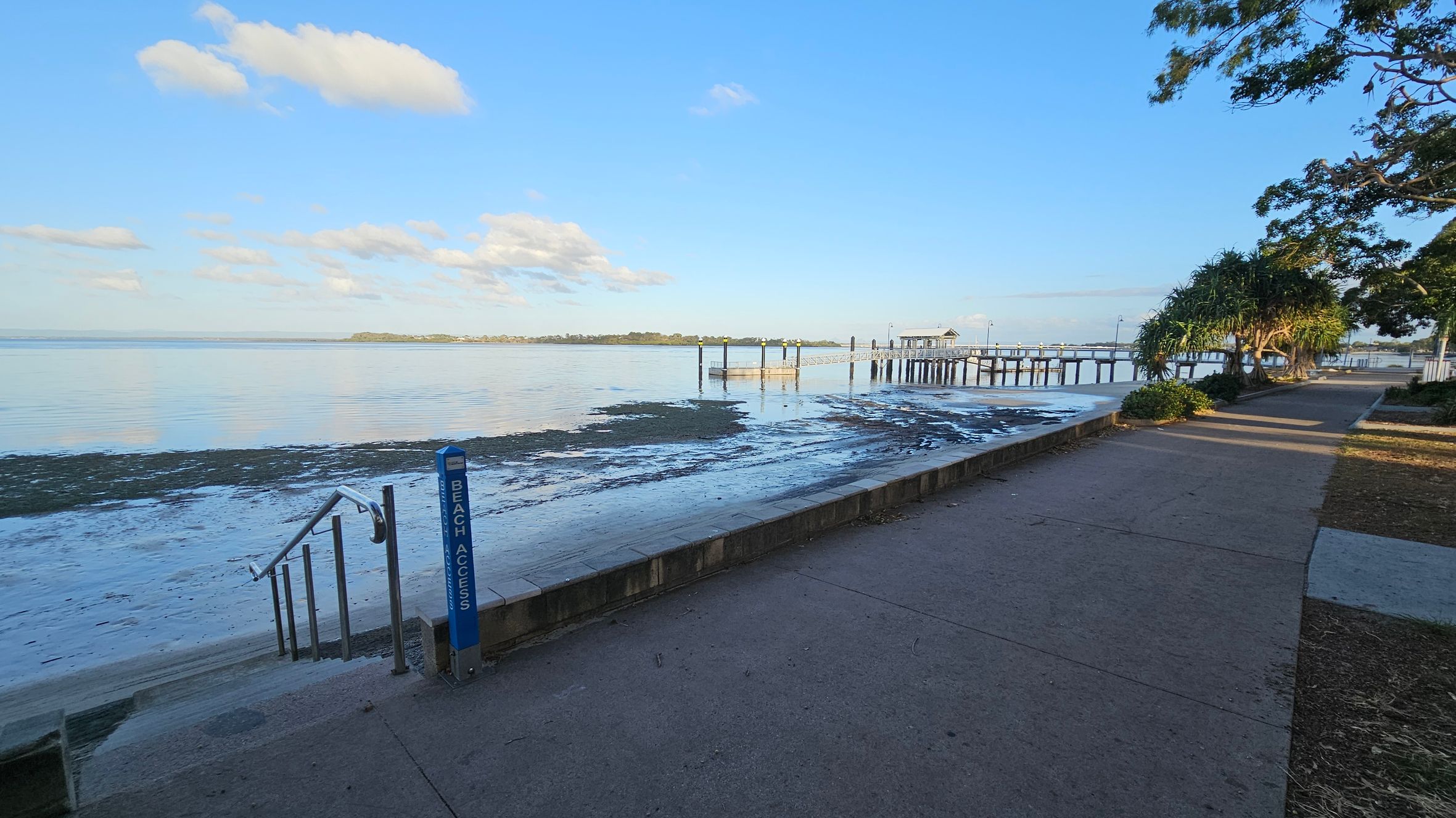 A coastal scene with a paved walkway on the right, bordered by trees and shrubs. A "Beach Access" sign and steps lead down to a sandy and muddy shoreline on the left. In the middle distance, a long pier extends over calm water, with a small structure at the end. The background features a distant landmass across the water. The sky is clear with a few clouds.