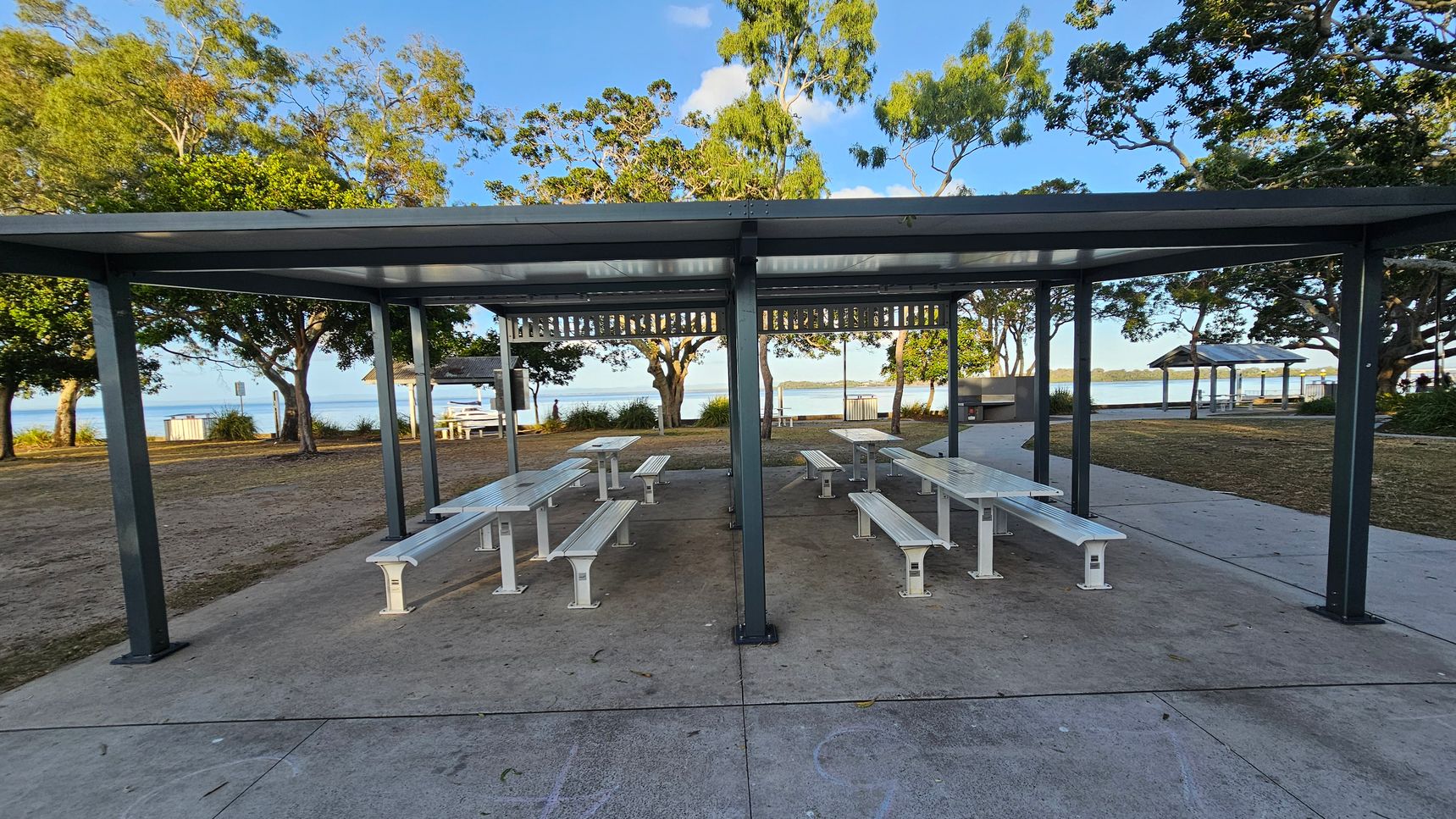 A covered picnic area with several white benches and tables on a concrete surface. The structure has a metal roof supported by dark green posts. In the background, there are trees with green foliage and a view of a body of water. To the right, a pathway leads to another covered area. The ground is a mix of grass and concrete, and the sky is clear.