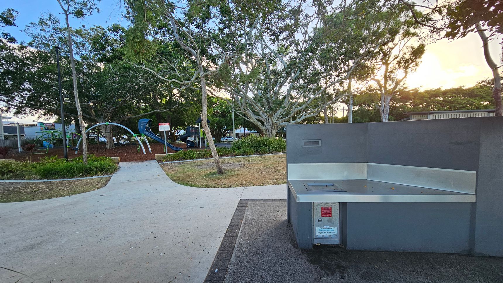 A park scene with a grey barbecue station on the right. In the middle, a paved path leads towards a playground area with climbing structures and slides. The left side features green grass and a variety of trees, including a large one with sprawling branches. The background is filled with more trees and a hint of a building. The ground is a mix of grass and mulch, and the sky is partially visible with a warm glow from the setting sun.