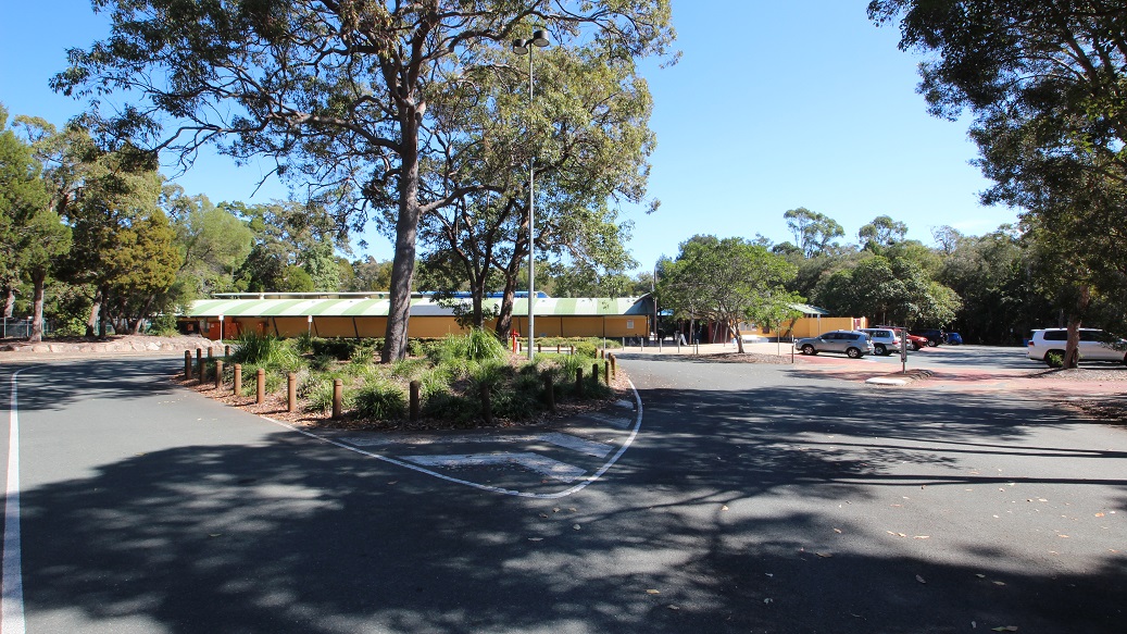 A plain driveway through the Bribie Island Aquatic and Leisure Centre. It is surrounded by beautiful bushland and forest, creating a serene atmosphere. You can spot a street light nearby, as well as two signs that probably look quite alike. The driveway is impeccably maintained, and the broken white lines along the road are clearly visible.