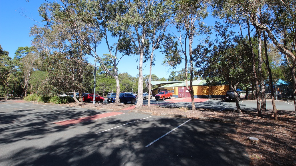 Another angle of the carpark inside the Bribie Island Aquatic and Leisure Centre. The area is filled with tall trees that provide a natural division between the two car spaces. It feels incredibly spacious and well-organized. In the background, you can spot a building painted mostly in yellow, with a striking red front gate.