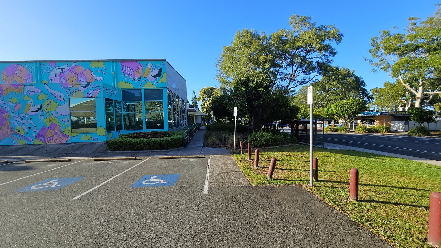 A colourful building on the left features a mural with whimsical designs, including fish and houses. Large windows reflect the surrounding greenery. In front, there are two marked disabled parking spaces. A pathway leads from the parking area towards a grassy section with bollards and a signpost. On the right, trees provide shade, and a road runs parallel to the building. A bus stop and more buildings are visible in the background.