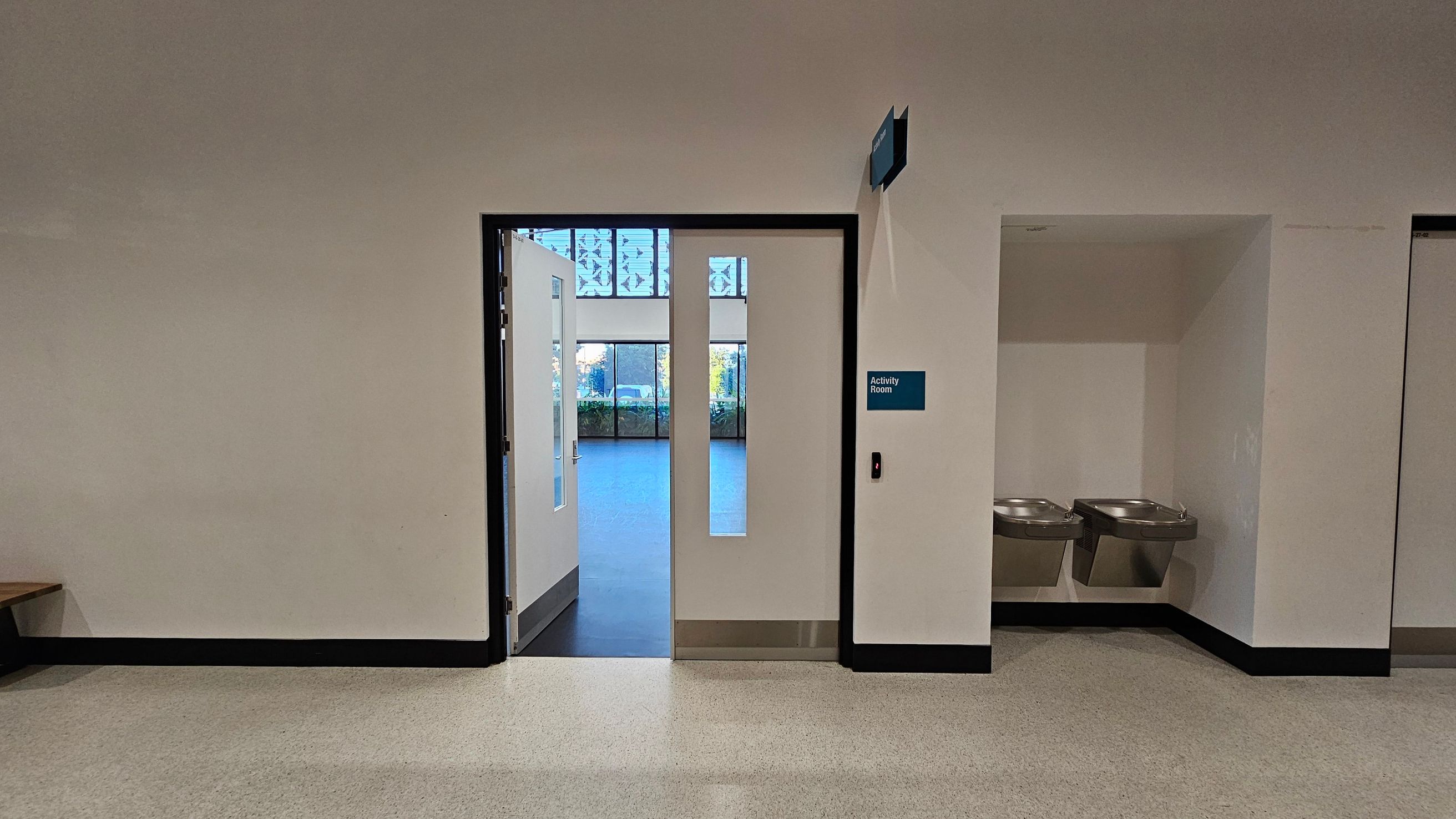 A hallway with a light-coloured floor and walls. In the centre, a double door is open, leading to a room with large windows showing greenery outside. To the right of the door, a sign reads "Activity Room" and there are two water fountains recessed into the wall. The ceiling is plain and light-coloured.