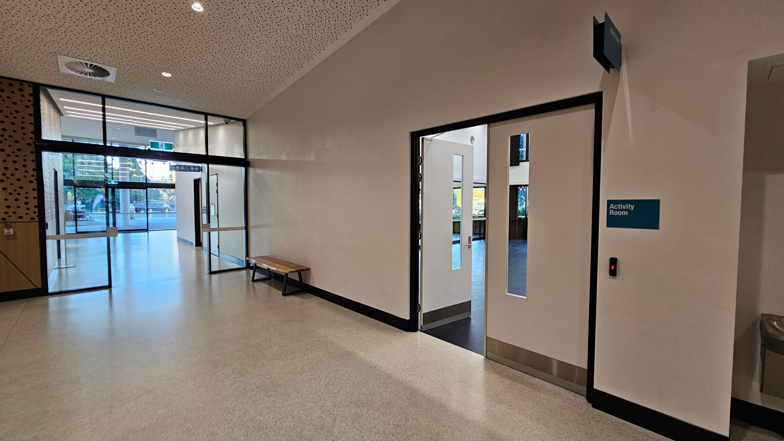 A hallway with a light-coloured floor and a perforated ceiling. On the right, there is a door labelled "Activity Room" with a small window. To the left, a glass entrance with double doors leads outside, with a wooden bench against the wall. The walls are plain, and there is a ventilation unit on the ceiling.