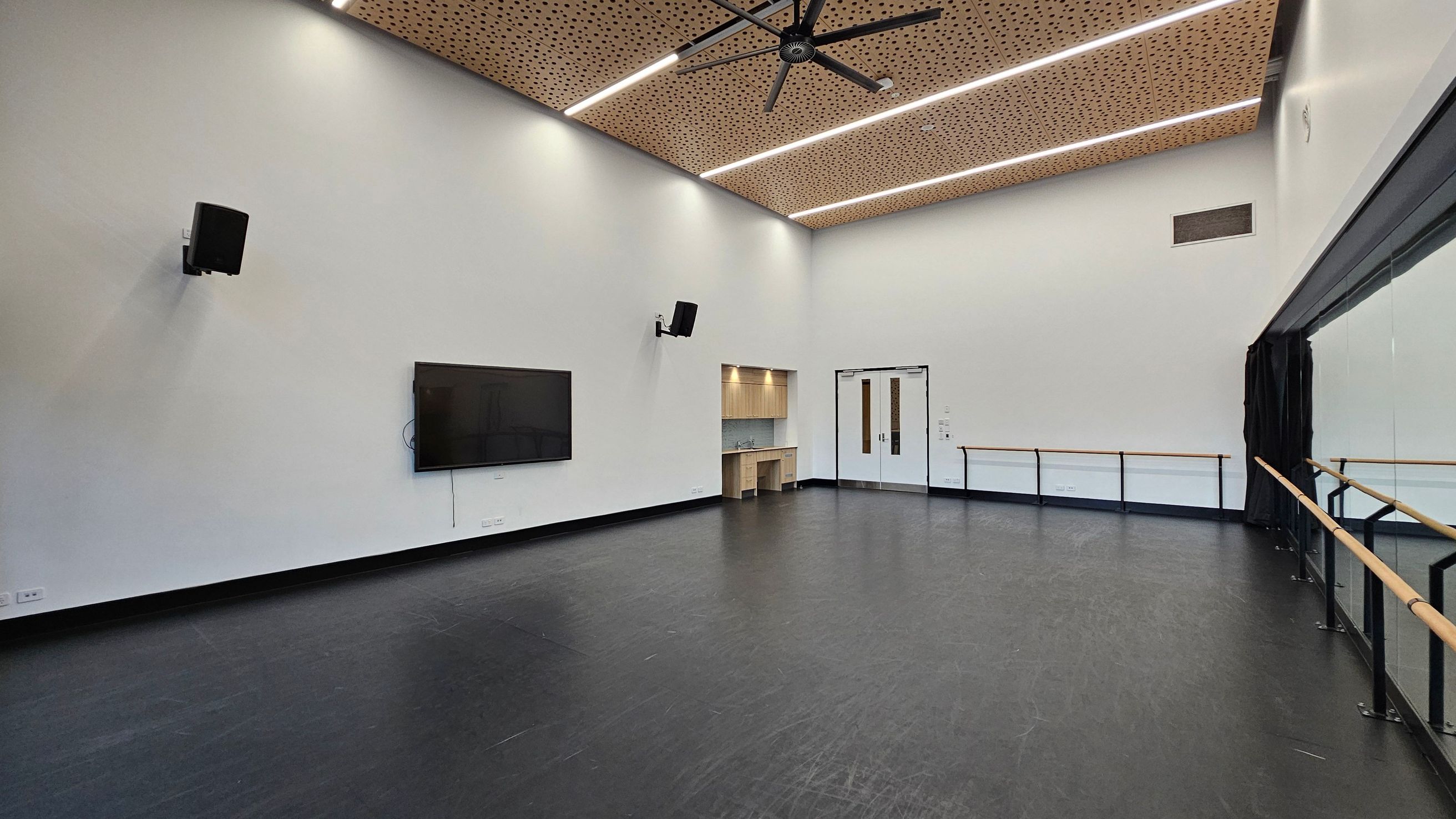 A spacious room with a black floor and white walls. On the left, a large flat-screen TV is mounted on the wall, flanked by two speakers. In the centre, there is a small kitchenette with a sink and wooden cabinets. The right side features a wall of mirrors with ballet barres. The ceiling is wooden with perforated panels and has a large fan and linear lights. Double doors are visible near the kitchenette.