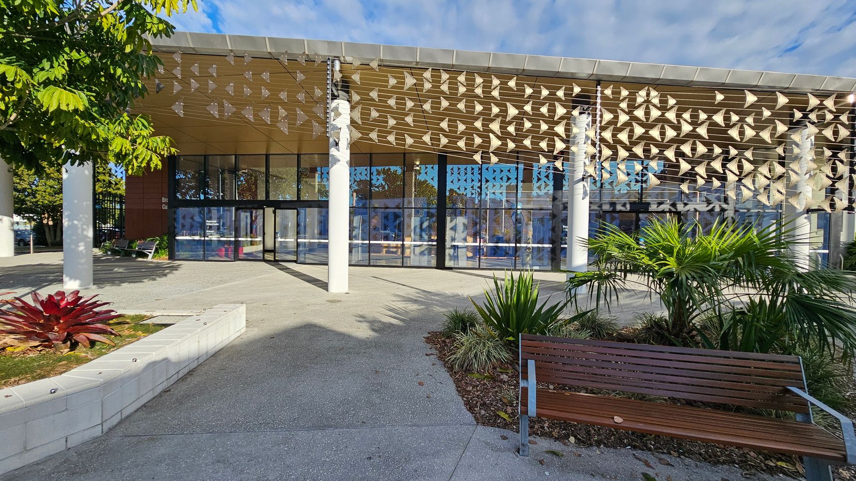 A modern building with a glass facade and a decorative overhang featuring geometric patterns. In the foreground, there is a wooden bench on the right and a landscaped area with various plants, including a red-leaved plant on the left. The ground is paved with concrete, and there are white columns supporting the structure. A tree with green leaves is partially visible on the left side.