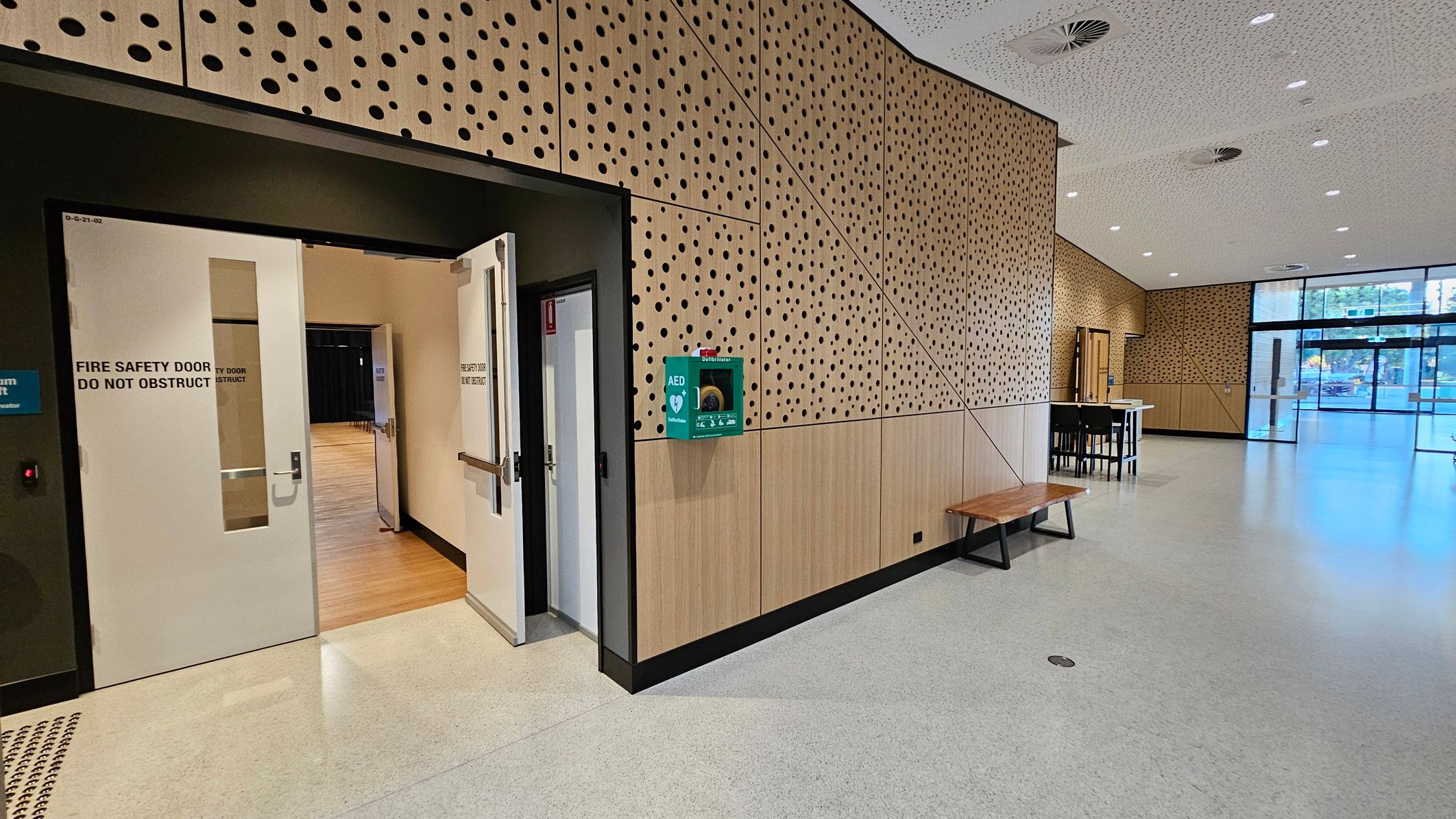 A spacious interior with a light-coloured, speckled floor and a perforated ceiling with circular vents. On the left, a fire safety door is slightly open, revealing a wooden floor inside. The wall is clad in wood panelling with circular cutouts, and an AED defibrillator is mounted on it. A wooden bench is positioned against the wall. On the right, large glass doors lead outside, and a small table with chairs is visible near the entrance.
