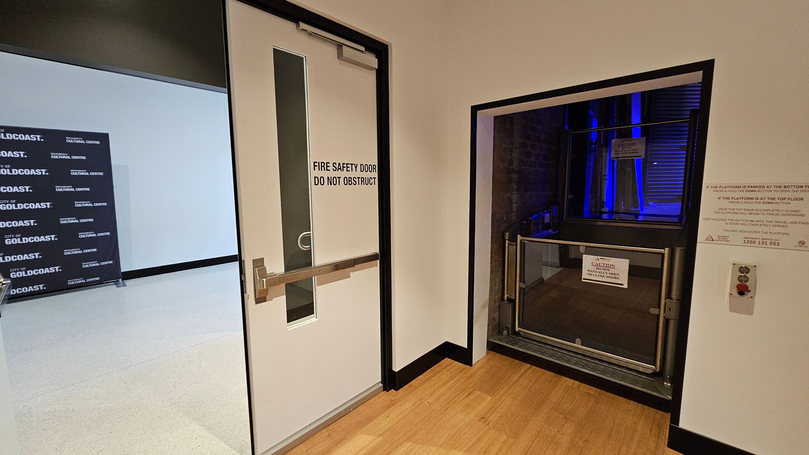 A white fire safety door with a vertical window and a push bar is on the left, marked "DO NOT OBSTRUCT." To the right, there's a platform lift with a caution sign, surrounded by metal railings. The lift area is illuminated with blue lighting. A control panel with buttons is mounted on the wall next to the lift. The floor is wooden, and the walls are light-coloured. In the background, a sign reads "CITY OF GOLDCOAST CULTURAL CENTRE."