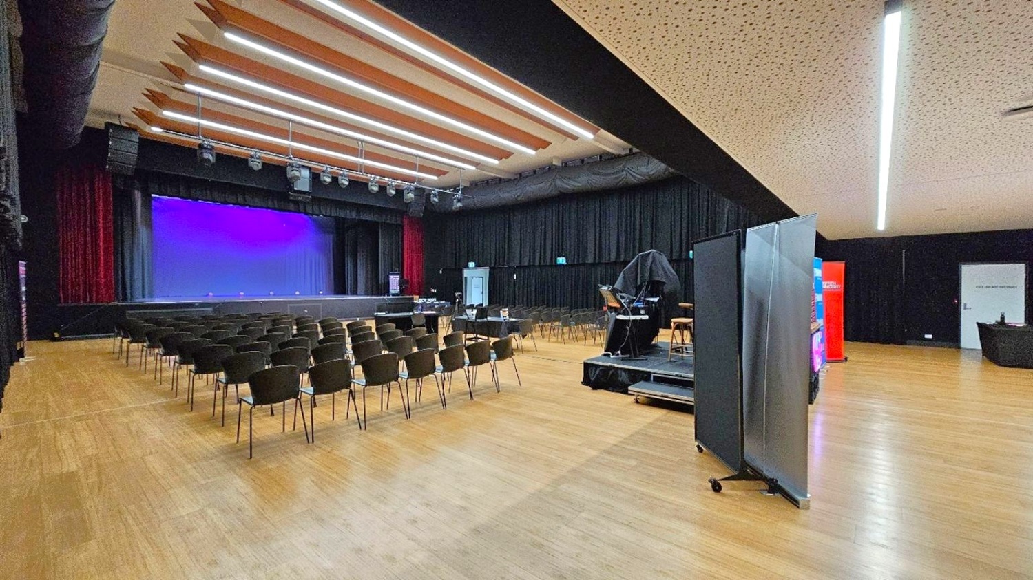 A spacious auditorium with a wooden floor and a perforated ceiling featuring linear lights. On the left, rows of black chairs face a stage. The stage is framed by red curtains. On the right, a covered keyboard and equipment are on a small platform. A folding screen and a red banner are nearby. The background has black drapes and a door marked "EXIT" on the right.