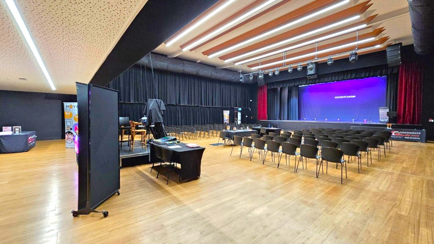 A spacious auditorium with a wooden floor and a ceiling featuring linear lights. On the left, there is a black partition and a table with promotional materials. In the centre, rows of black chairs face a stage. The stage has a large purple backdrop. On the right, a table with more promotional items is visible. The stage is equipped with lighting and sound equipment.