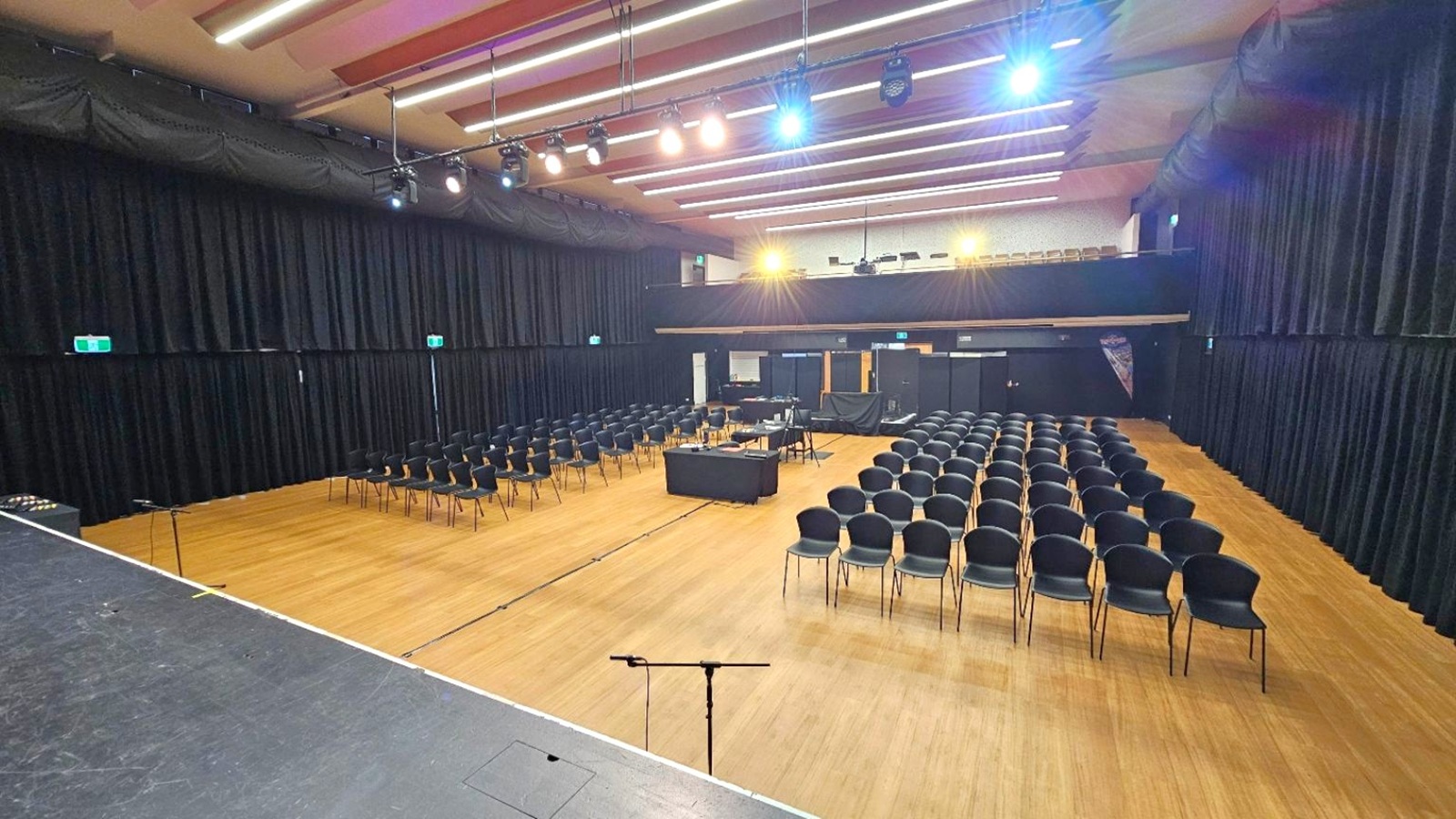 A large auditorium with a wooden floor and a high ceiling featuring rows of lights. On the left, there is a stage with a microphone stand. In the centre, rows of black chairs are arranged facing the stage, with a table in the middle. The background has a control area with equipment and a banner. Black curtains line the walls, and there are exit signs visible. The lighting is bright, illuminating the entire space.