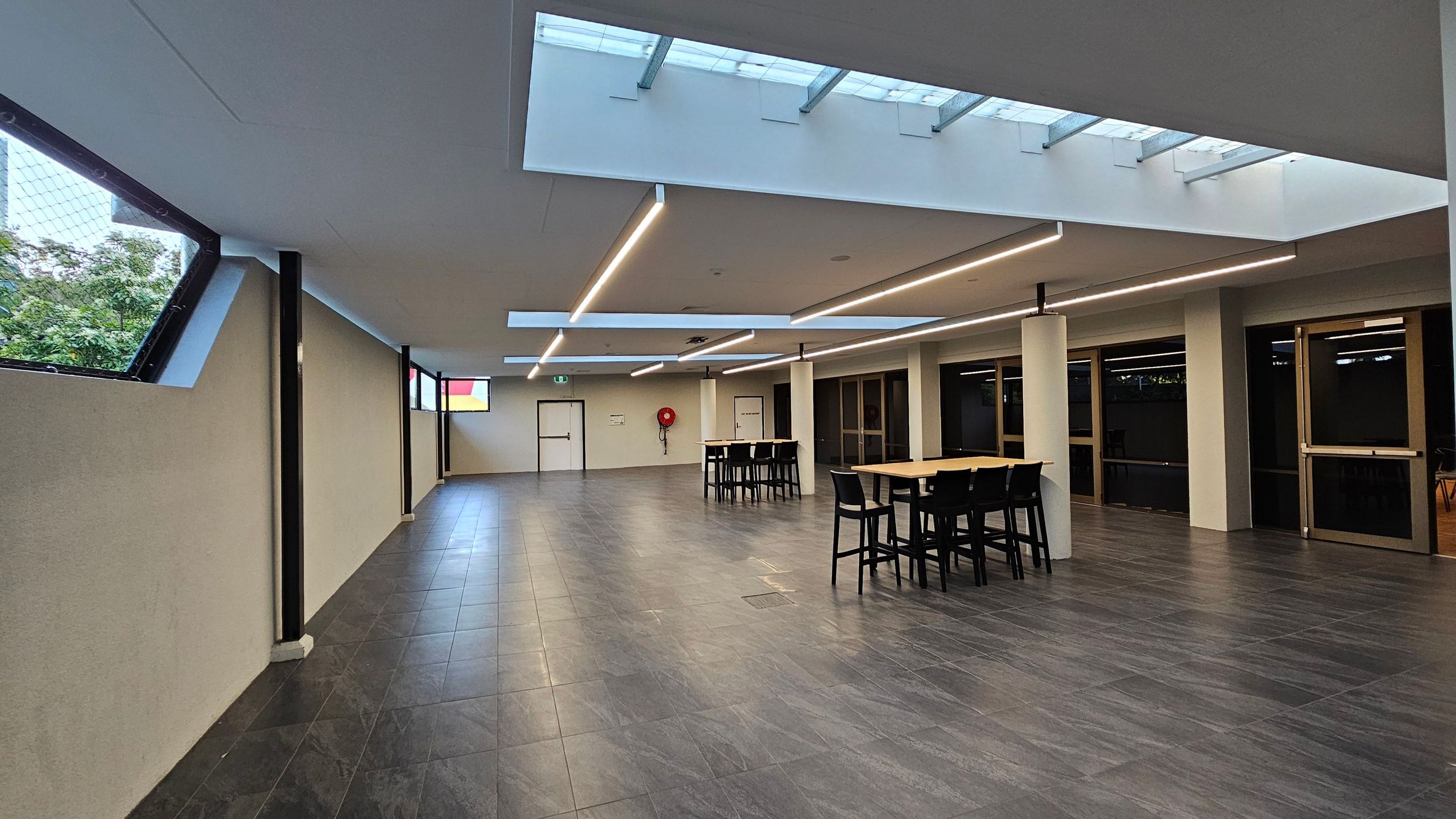 A spacious indoor area with a grey tiled floor and a ceiling featuring skylights. On the right, there are several tall tables with black chairs. The left side has slanted windows with a view of greenery outside. The back wall includes a door and a fire hose reel. The ceiling has modern linear lighting fixtures.