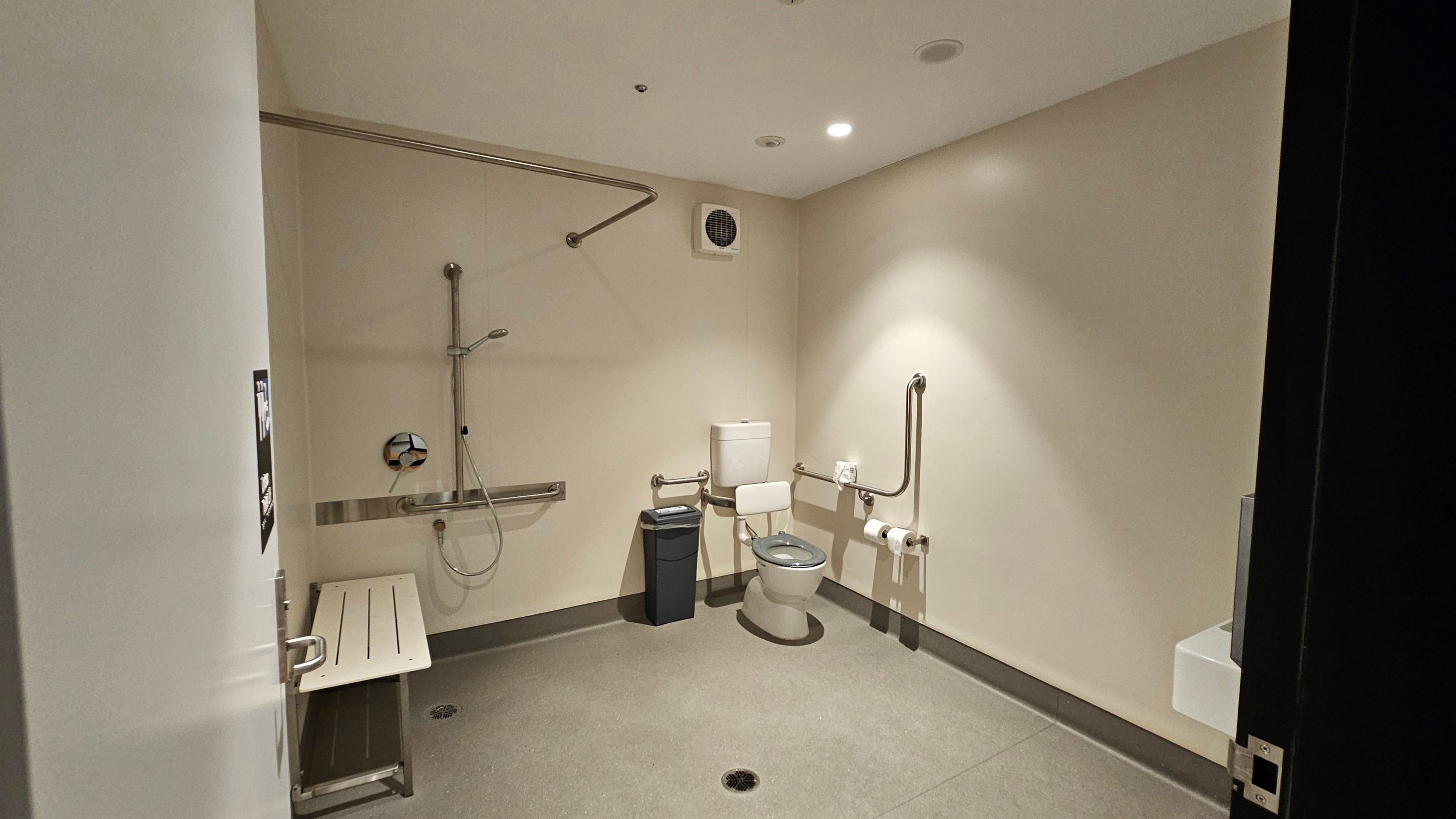 Accessible bathroom with a shower area on the left, featuring a wall-mounted showerhead and a foldable bench. In the centre, a toilet with support rails on both sides and a bin nearby. Toilet paper holders are mounted on the right wall. The floor is non-slip, and the ceiling has recessed lighting. A ventilation fan is visible above the toilet.