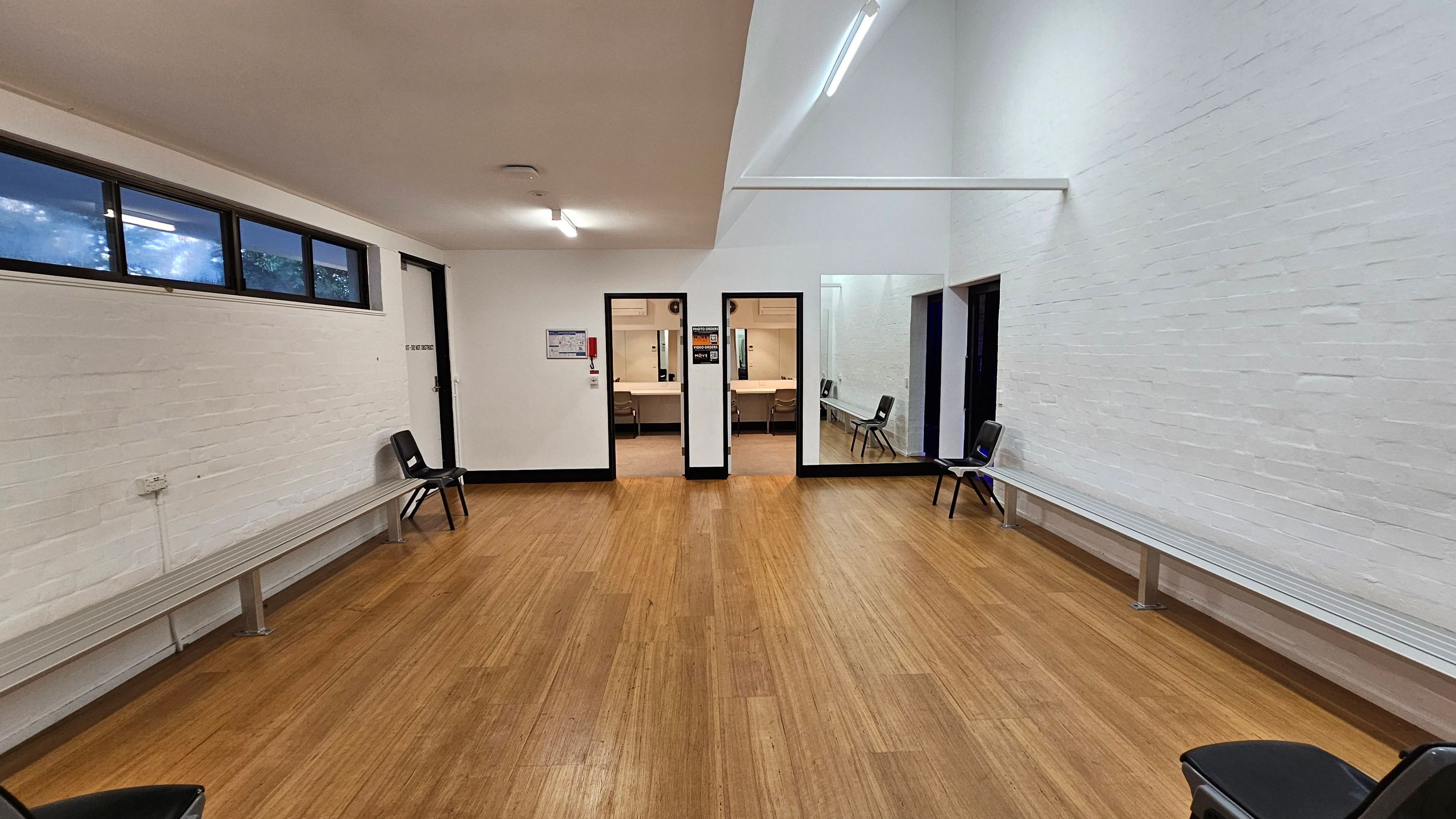 A room with wooden flooring and white brick walls. On the left, there are high windows and a long bench with a few black chairs. In the centre, two doorways lead to another room with tables and chairs. On the right, more black chairs are placed along the wall. The ceiling is white with fluorescent lights.