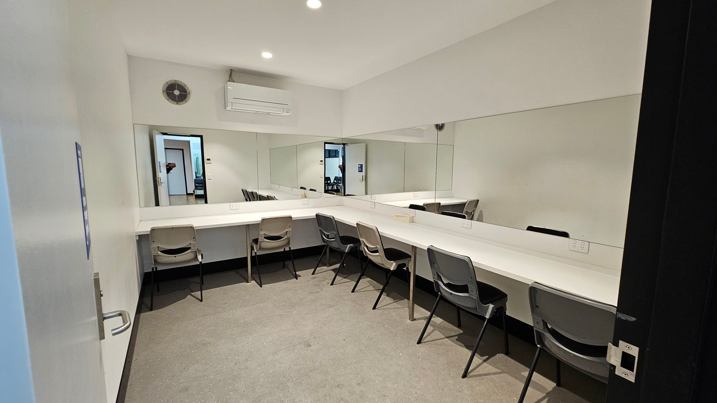 A room with a long white counter along the right wall, featuring several grey chairs. A large mirror spans the wall above the counter. On the left wall, there's a clock and an air conditioning unit. The floor is carpeted in a neutral tone. A door on the left leads to another room, visible through the mirror's reflection. The ceiling is plain with recessed lighting.
